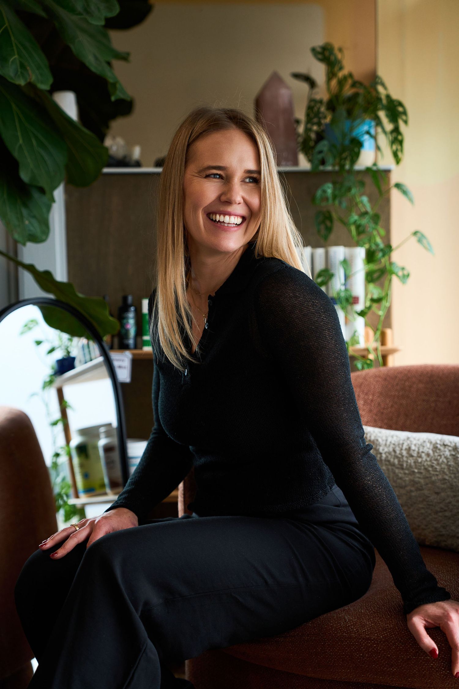 Smiling blonde woman in black turtleneck sits in a modern salon surrounded by lush green plants.