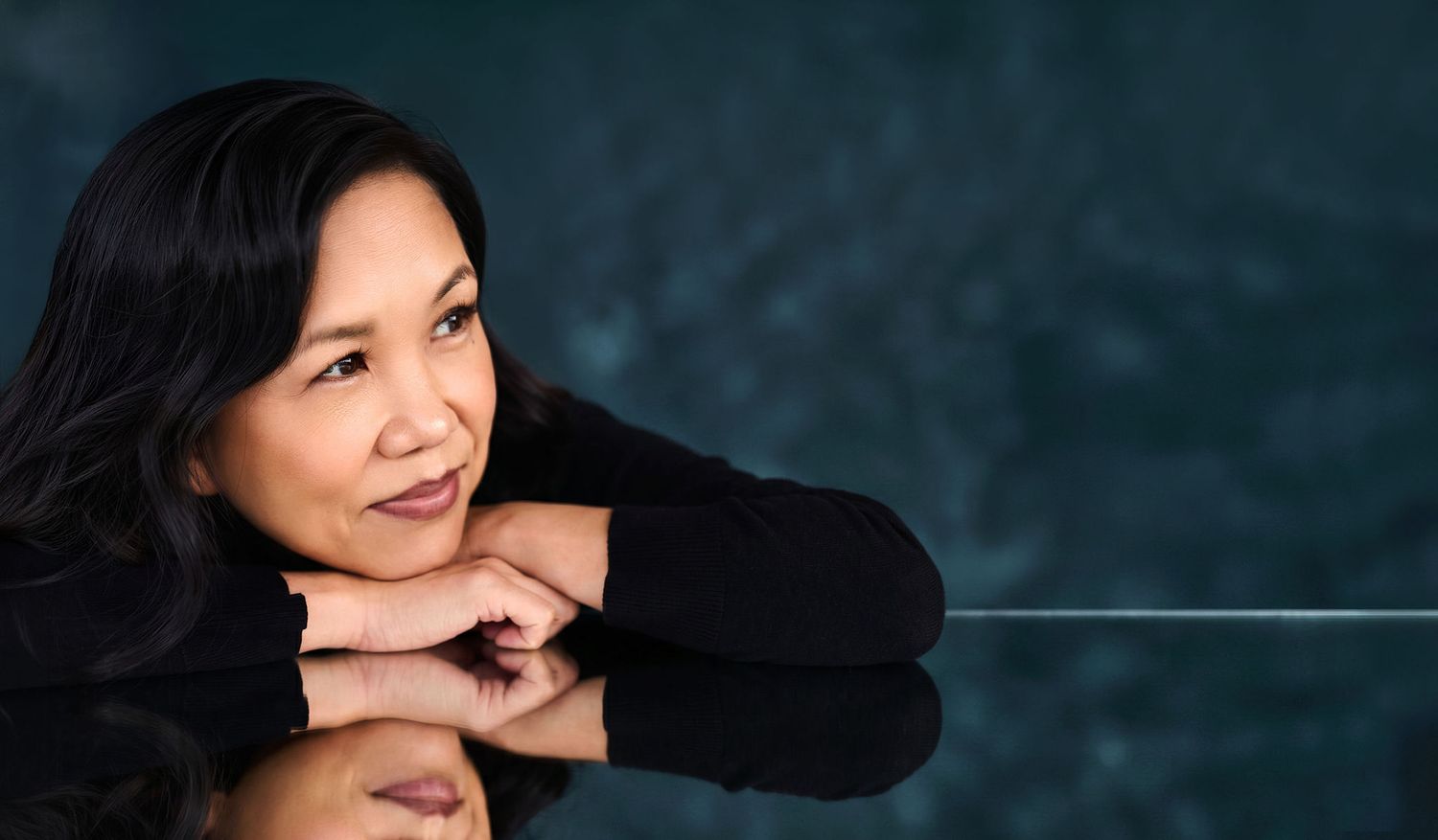 A woman with long black hair rests her chin on her hands, posing thoughtfully against a dark teal background.