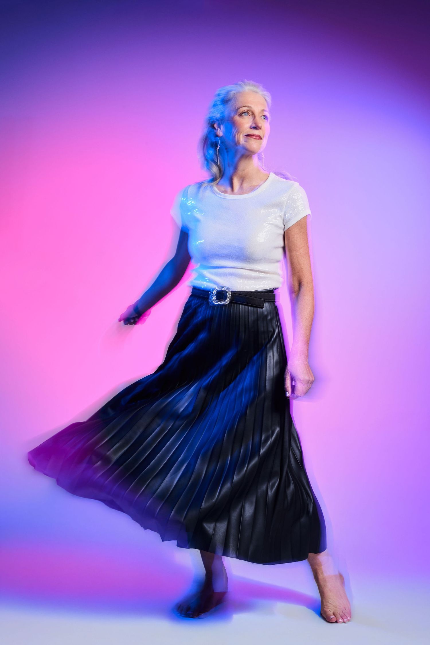 Elegant older woman with silver hair posing in white tee and black pleated skirt under vibrant pink and purple studio lighting.
