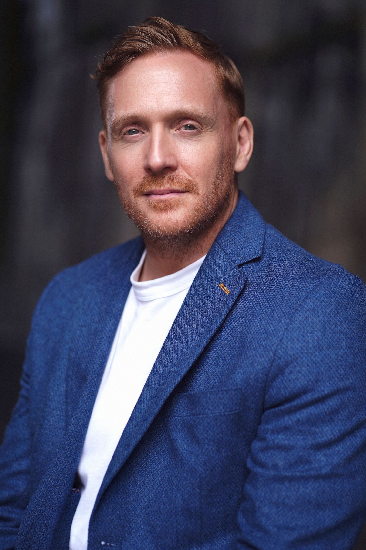 A person wearing a navy blue blazer and white t-shirt poses for a professional headshot against a dark background. Studio TingTing.