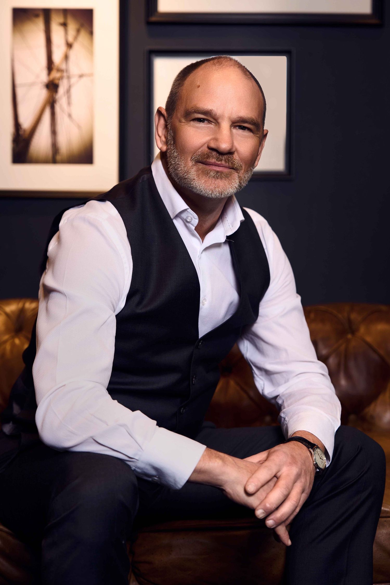 Professional headshot of a person in a black vest and white shirt seated on a brown leather couch in a Sydney studio setting.