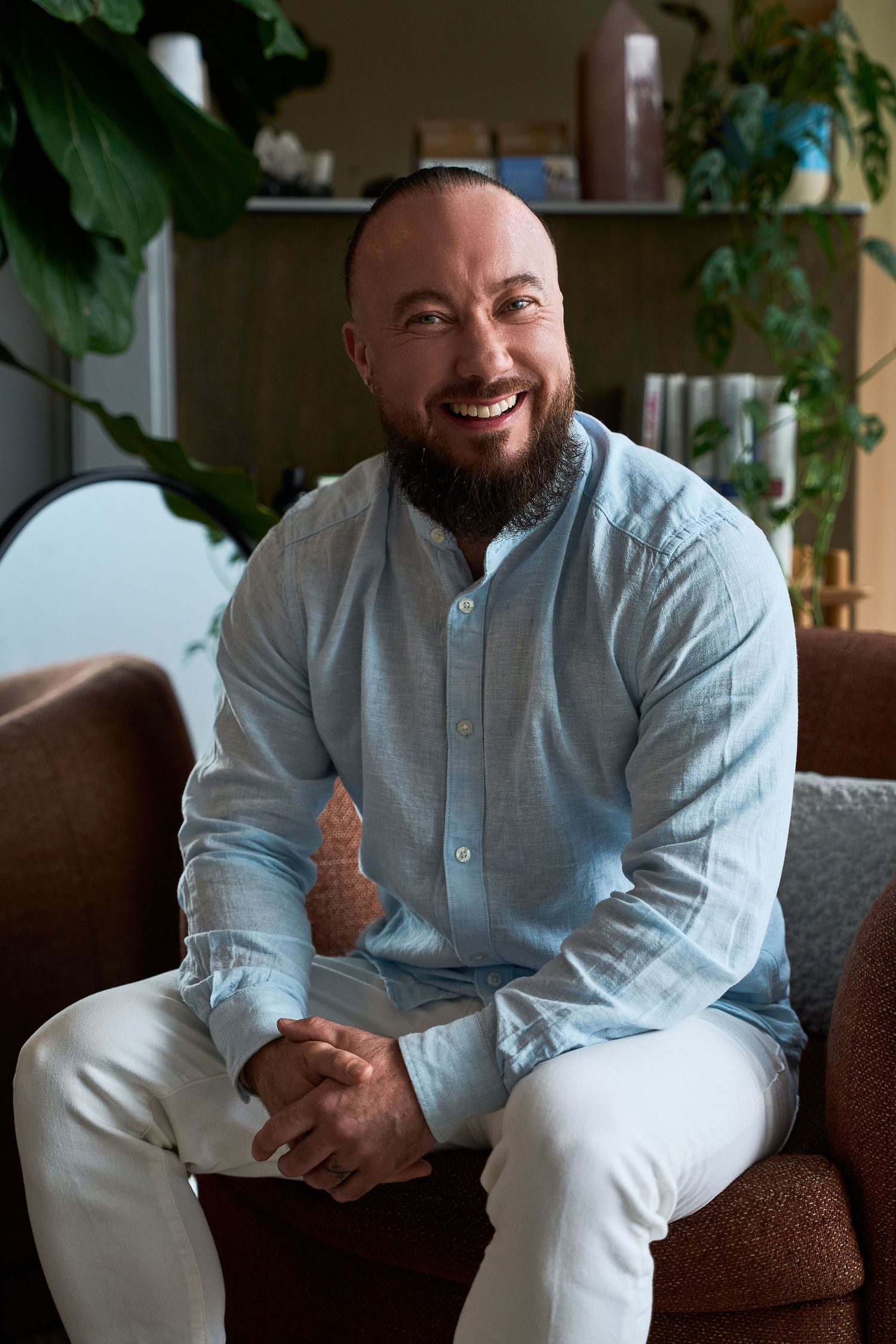Smiling bearded bald man in light blue linen shirt and white pants sitting on brown leather sofa indoors.