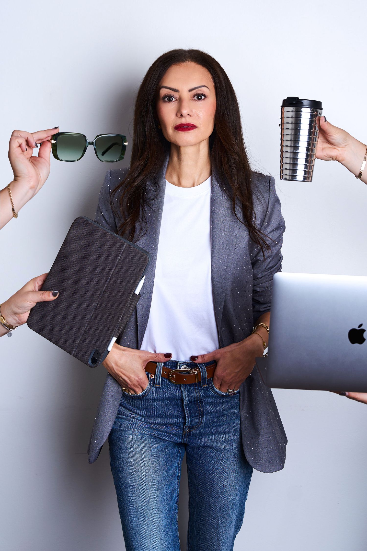 Busy businesswoman in gray blazer and jeans surrounded by hands offering sunglasses, coffee, tablet, and laptop.
