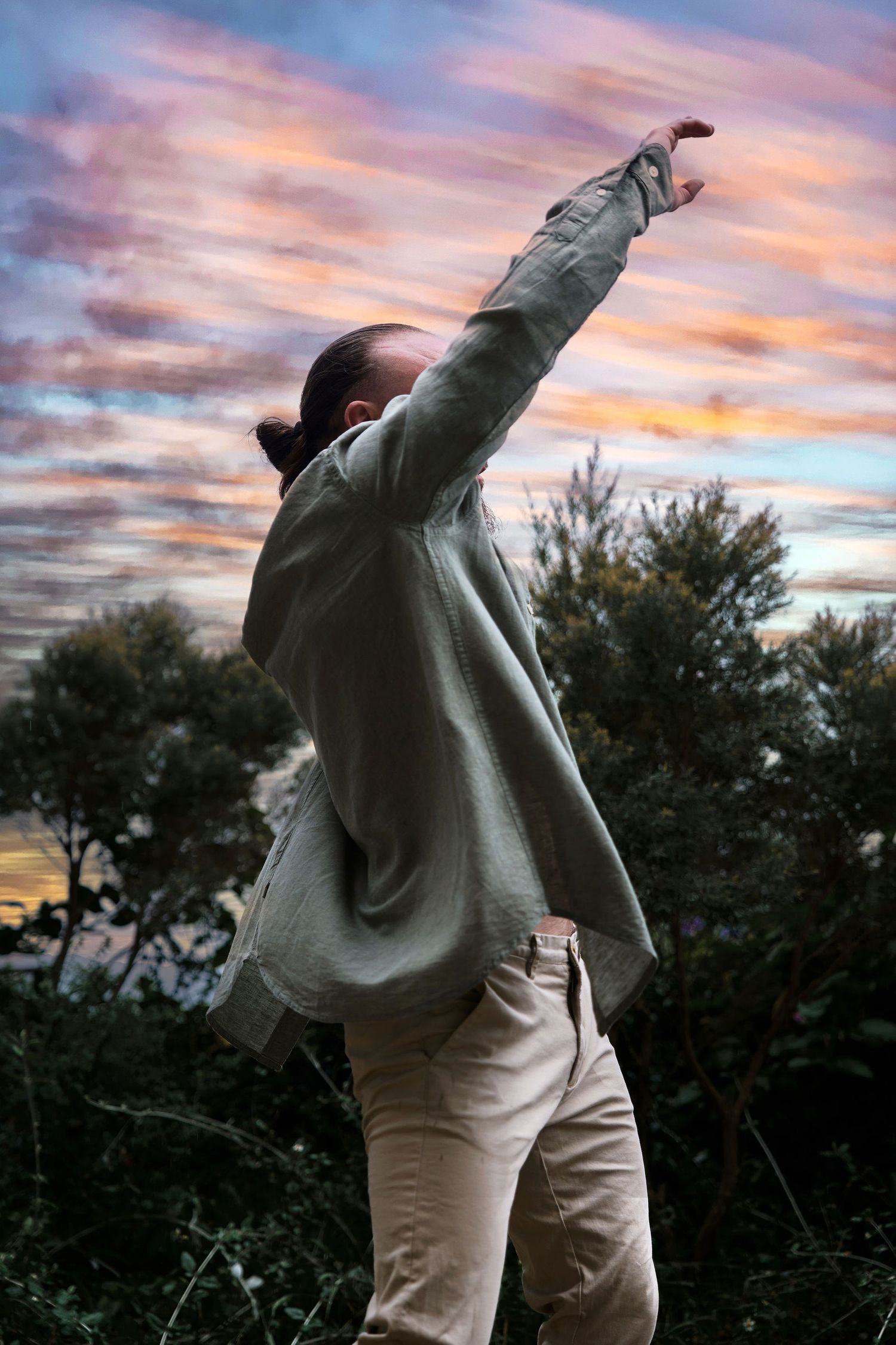 Person with arm raised against a vibrant sunset sky with colorful clouds and trees in background.