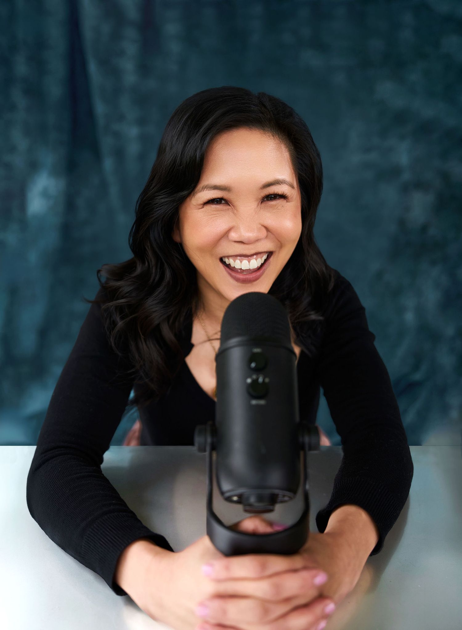 Smiling woman with dark wavy hair poses with a professional microphone at a table against a teal backdrop.