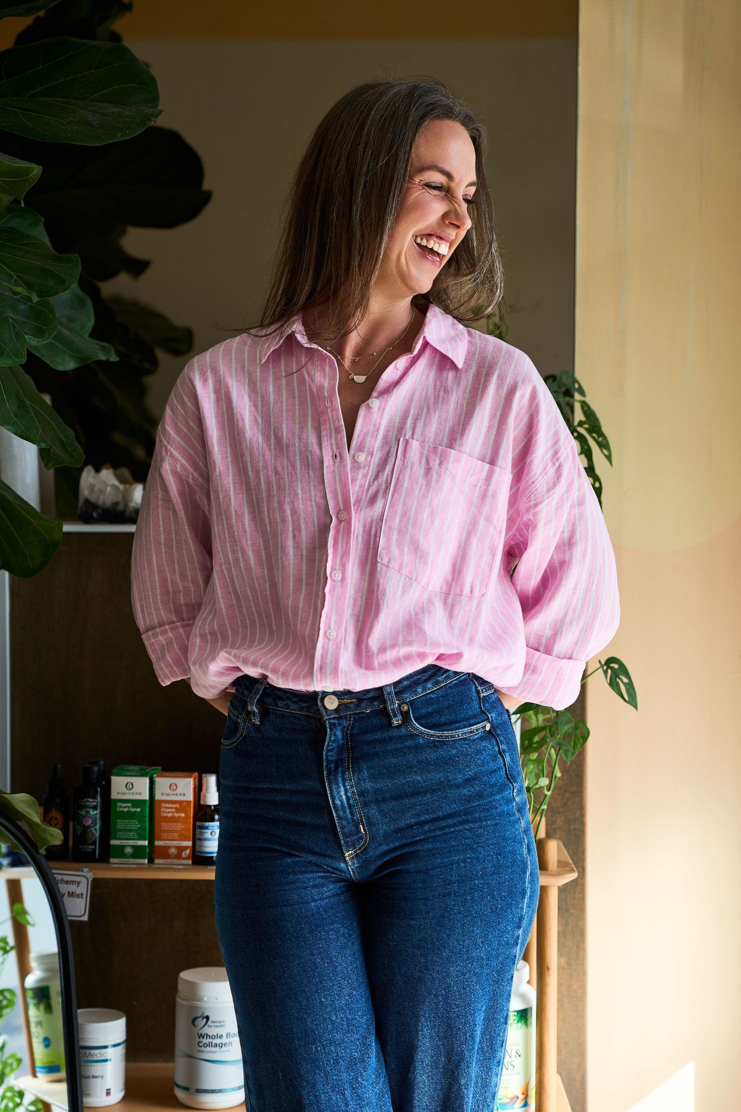 Smiling woman in pink striped shirt and blue jeans standing in a bright room with green plants and supplements.