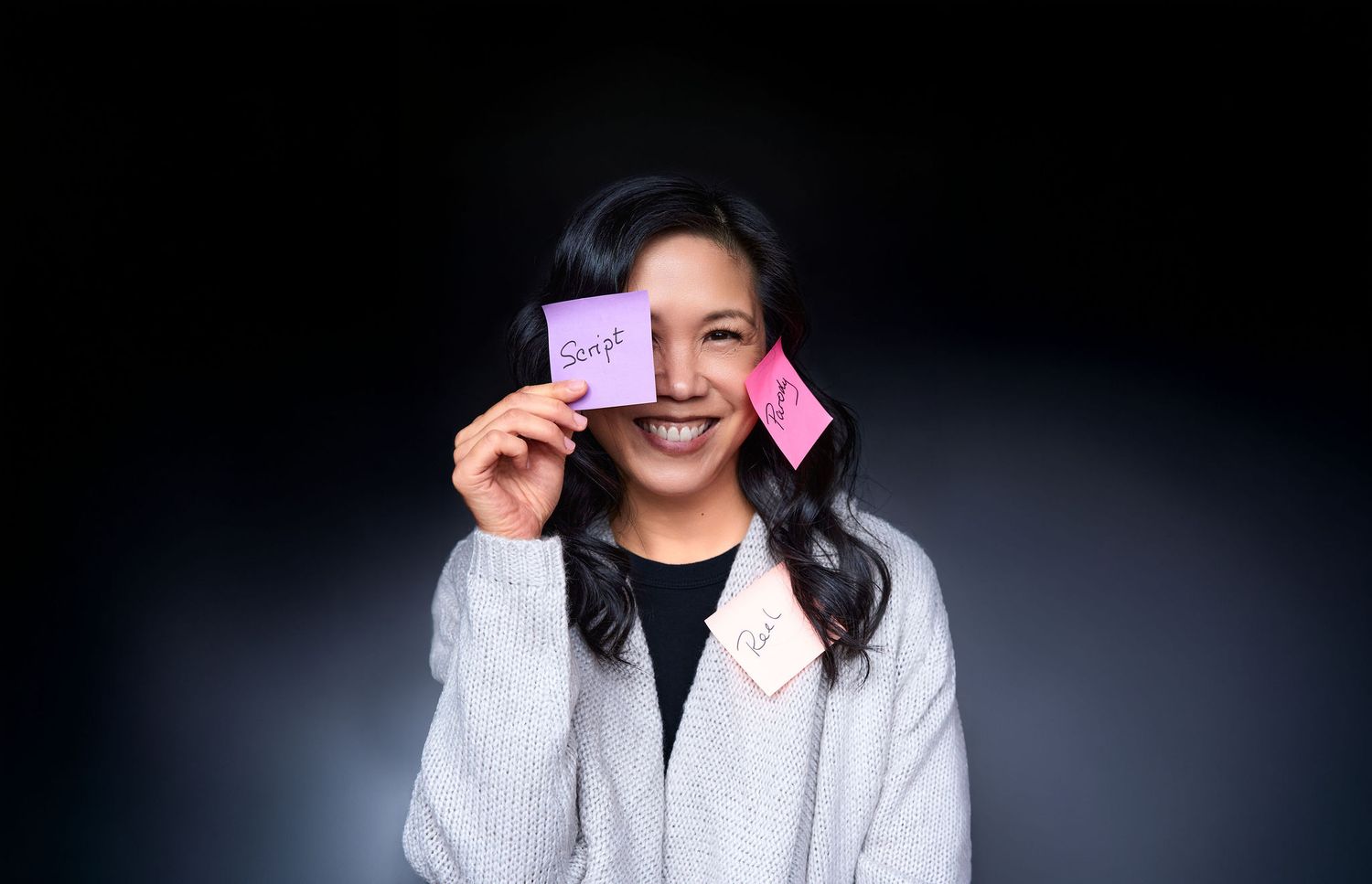 Smiling young woman holding colorful sticky notes over her eyes against a dark background.