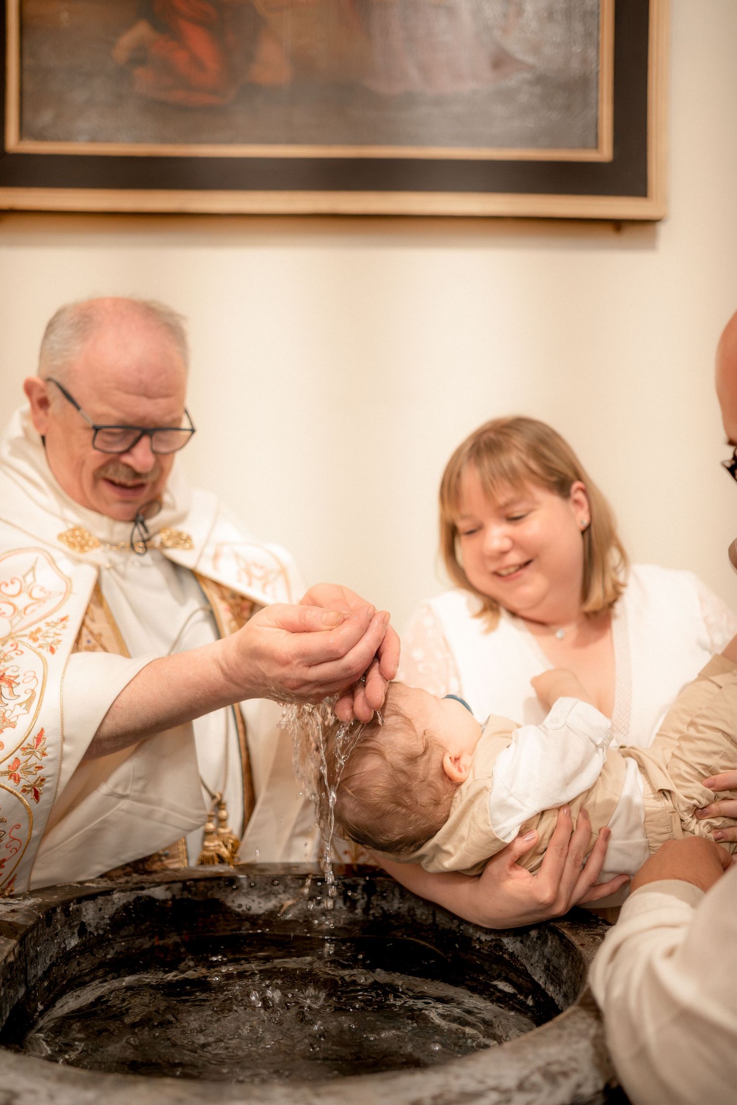 Priest performing Catholic baptism ceremony, pouring holy water over infant held by smiling mother at baptismal font.