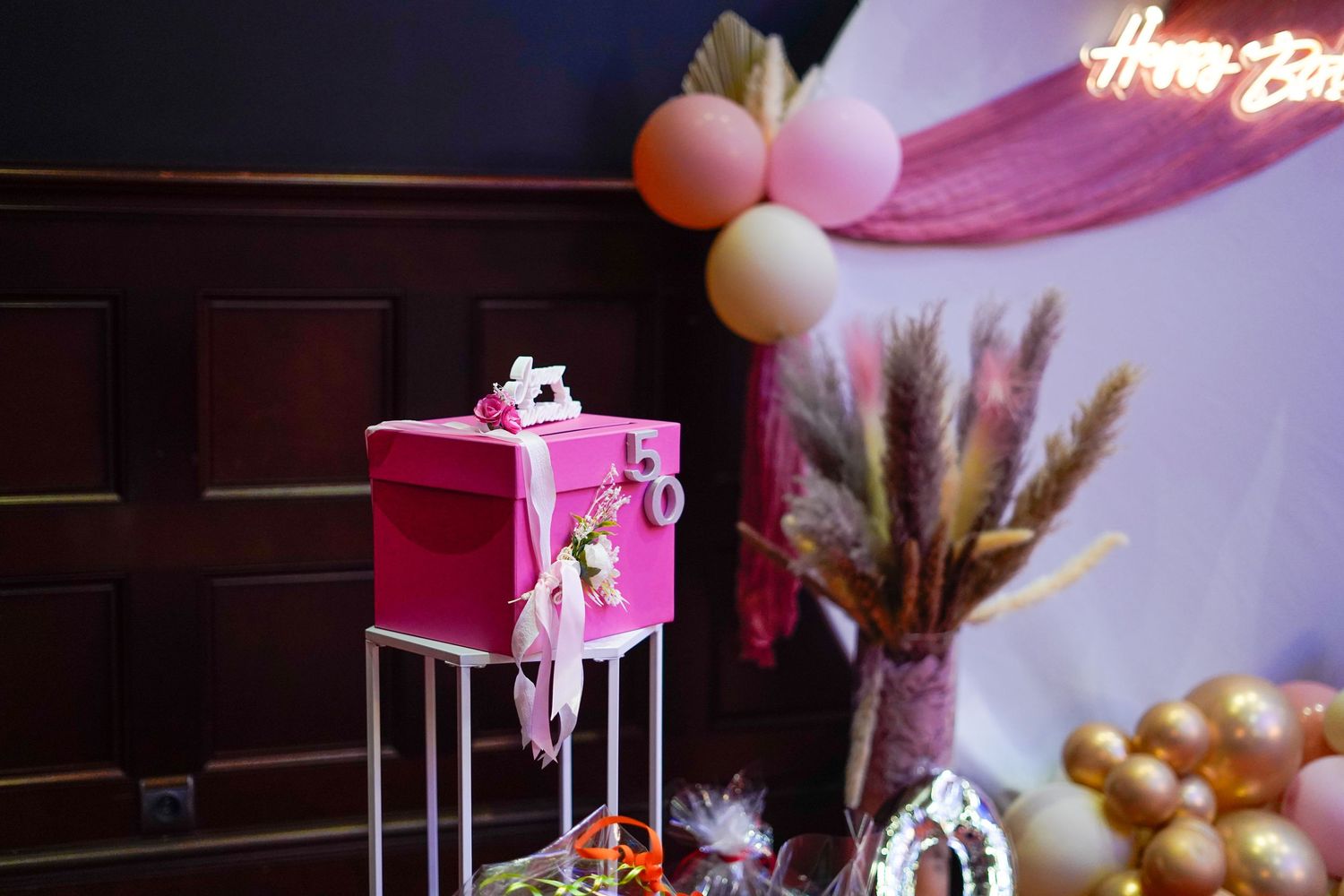 Pink gift box on stand with Happy Birthday neon sign, balloons, and dried flowers in festive party decoration setup.