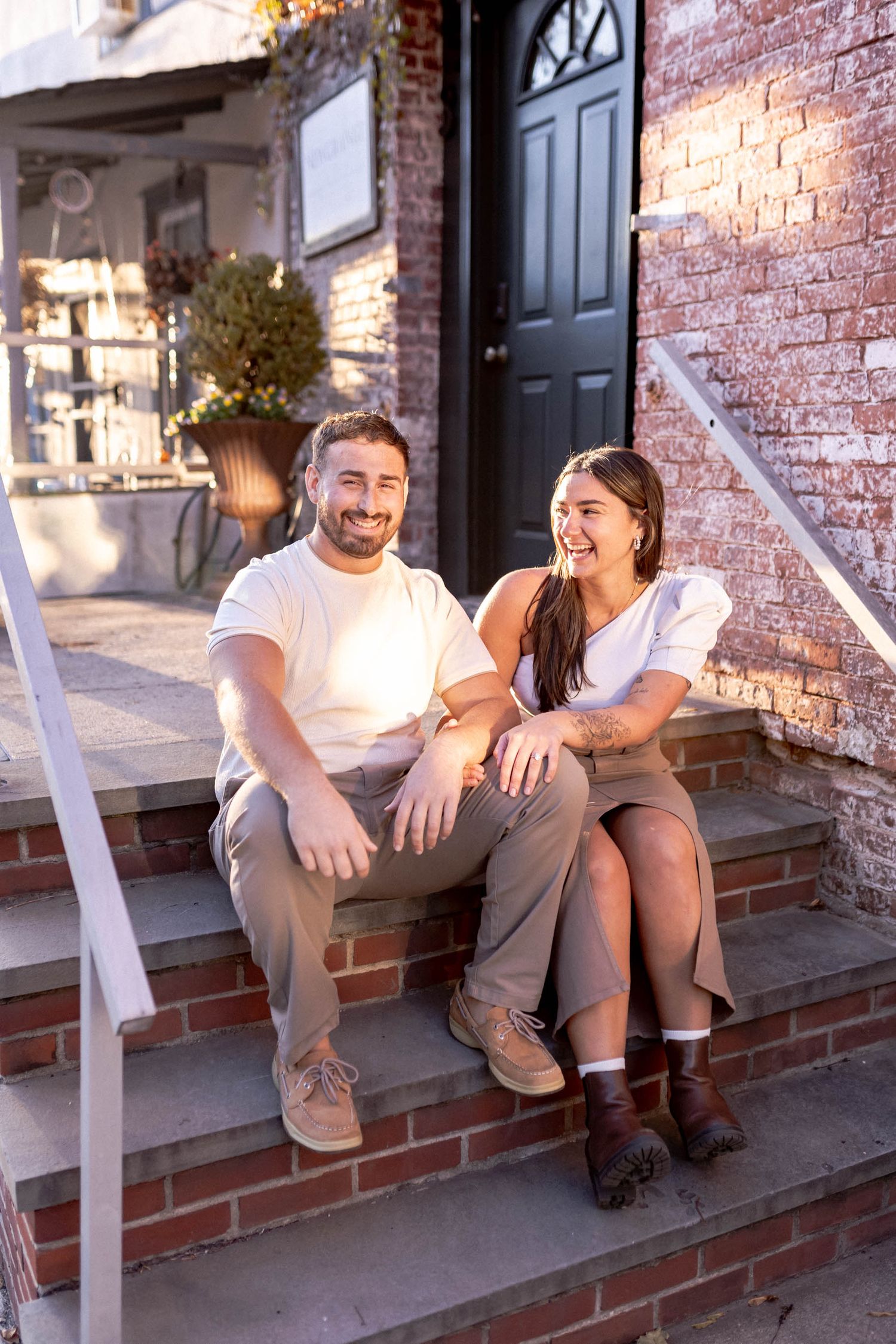 A couple sitting and laughing together on brick steps outside a black door on a sunny day.
