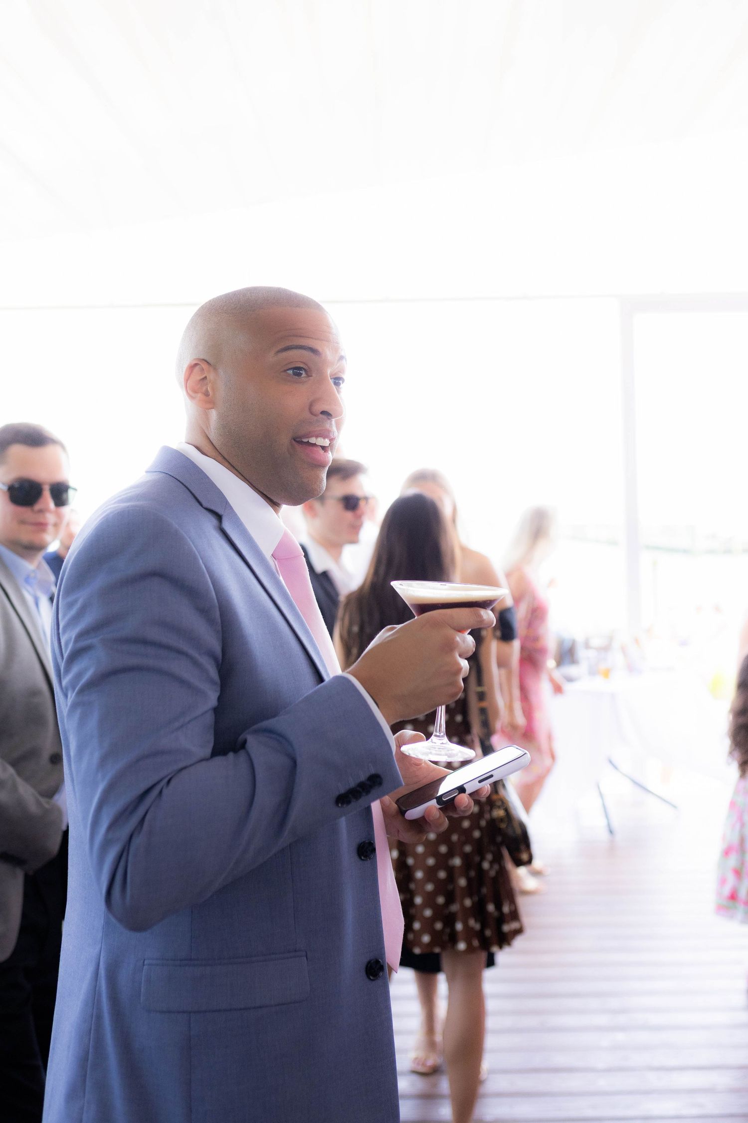 A man in a blue suit jacket gestures while speaking at an event with other guests visible in the background.