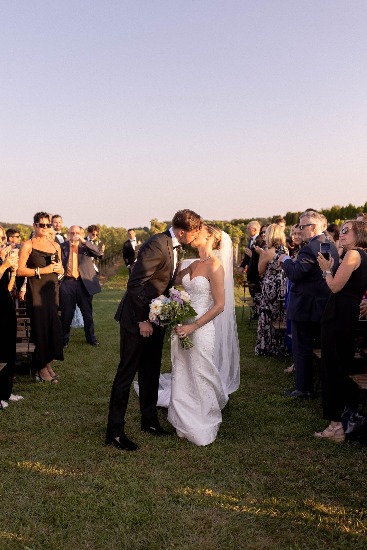 Wedding party and guests mingling on a grassy field during sunset celebrations.
