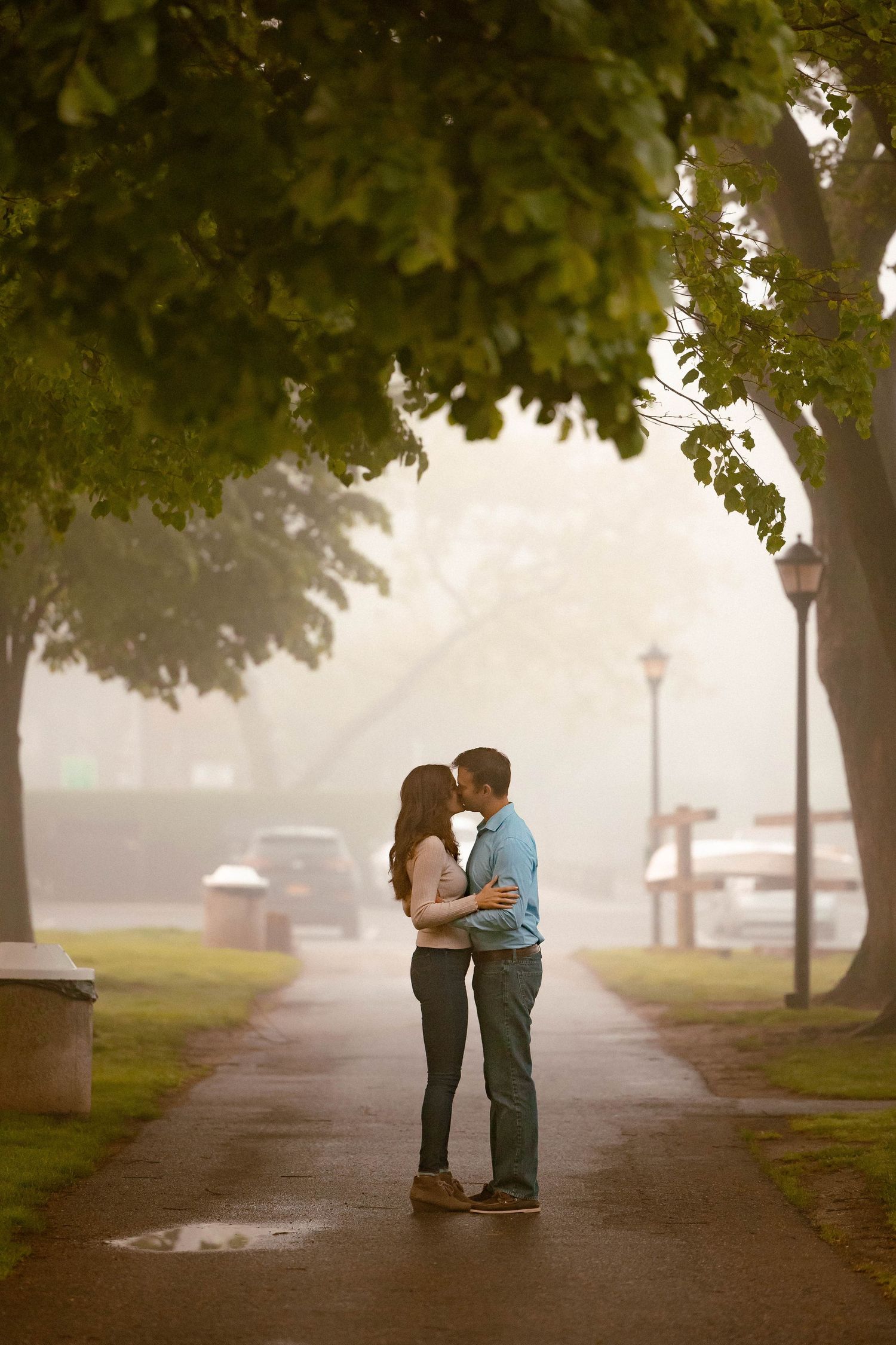 A romantic couple embraces on a misty tree-lined path during a foggy morning.