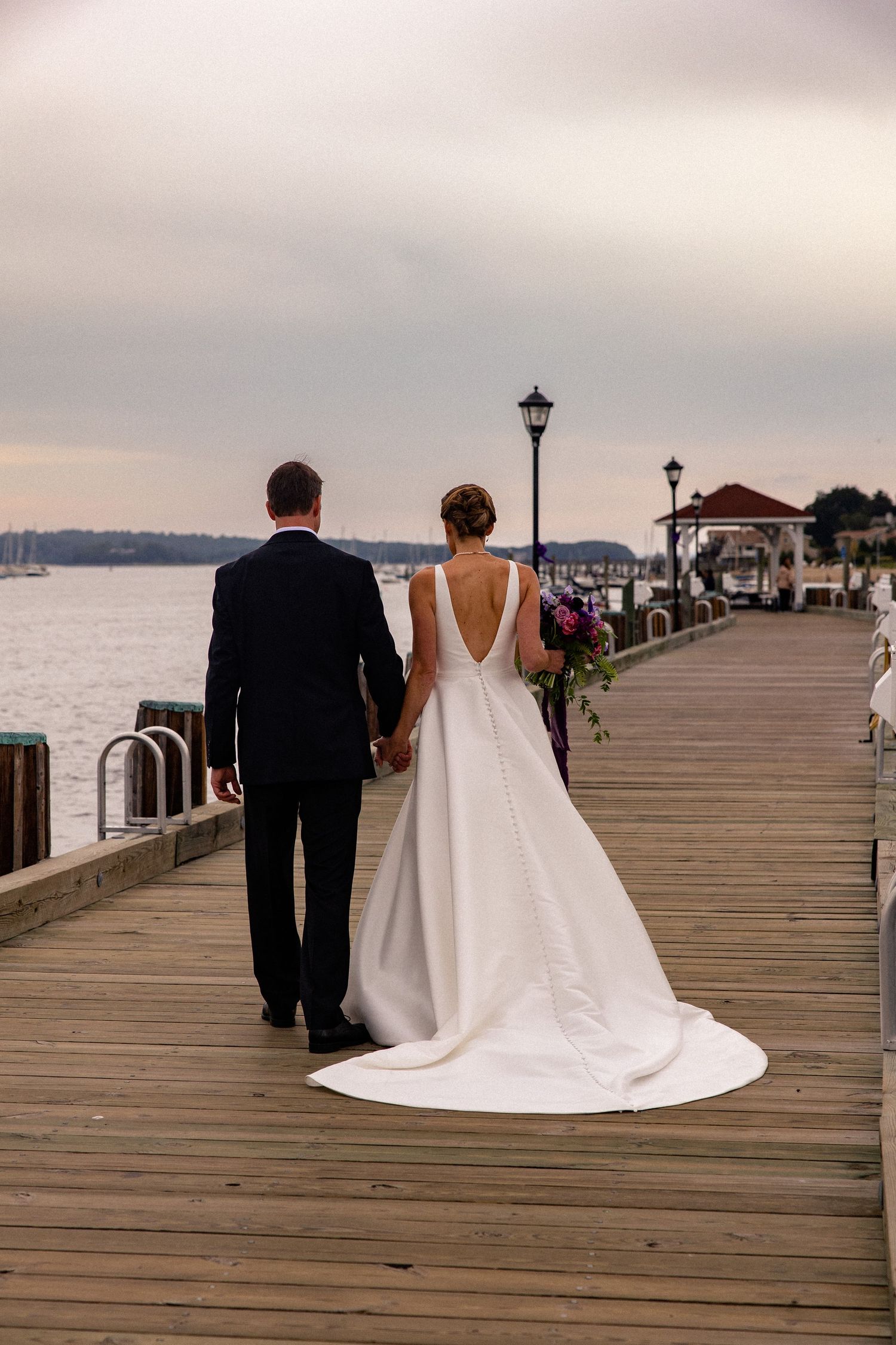 A couple in wedding attire walk along a waterfront boardwalk with a gazebo in the distance at sunset.