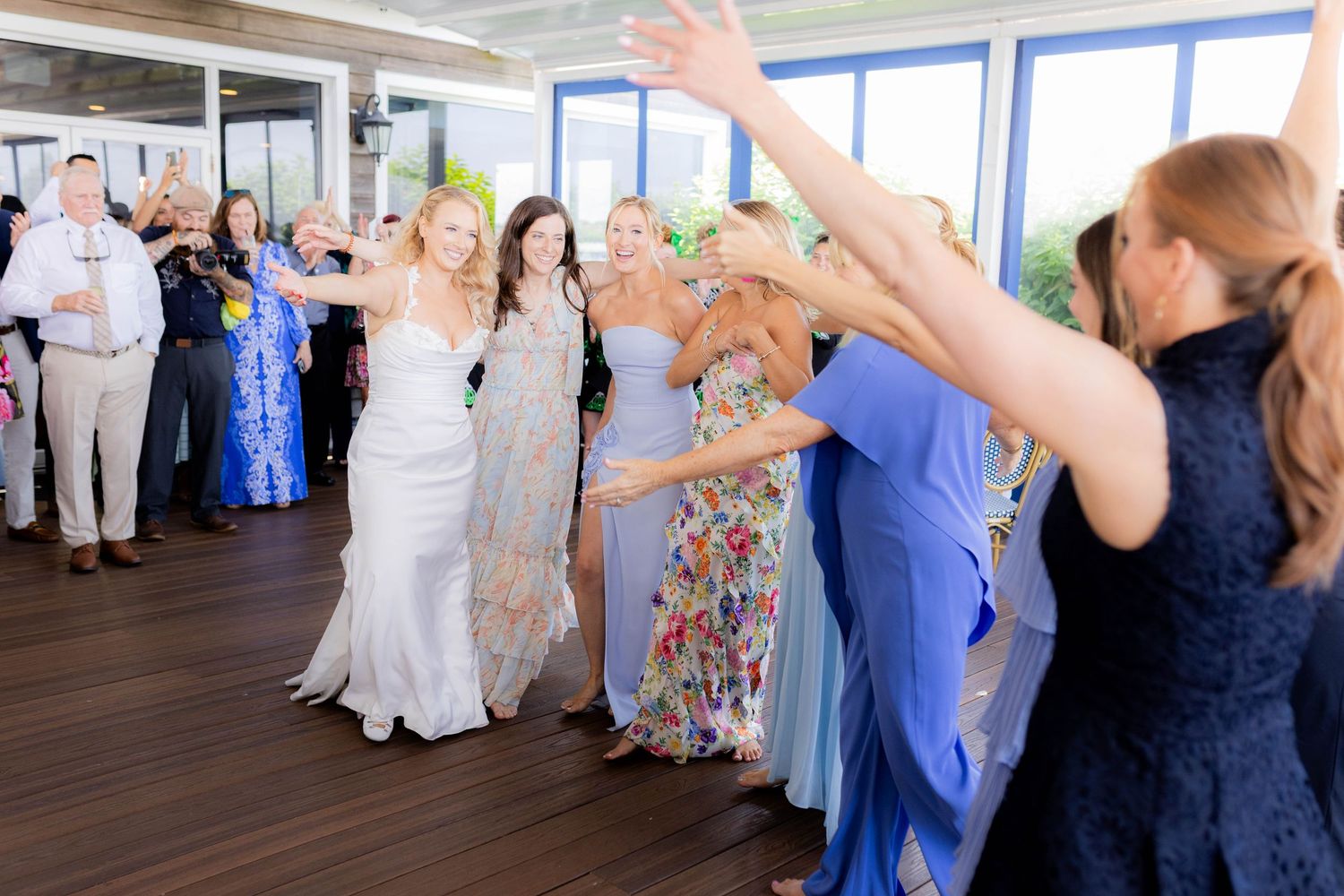 Wedding guests dance and celebrate together on a wooden dance floor at a reception venue with large windows.