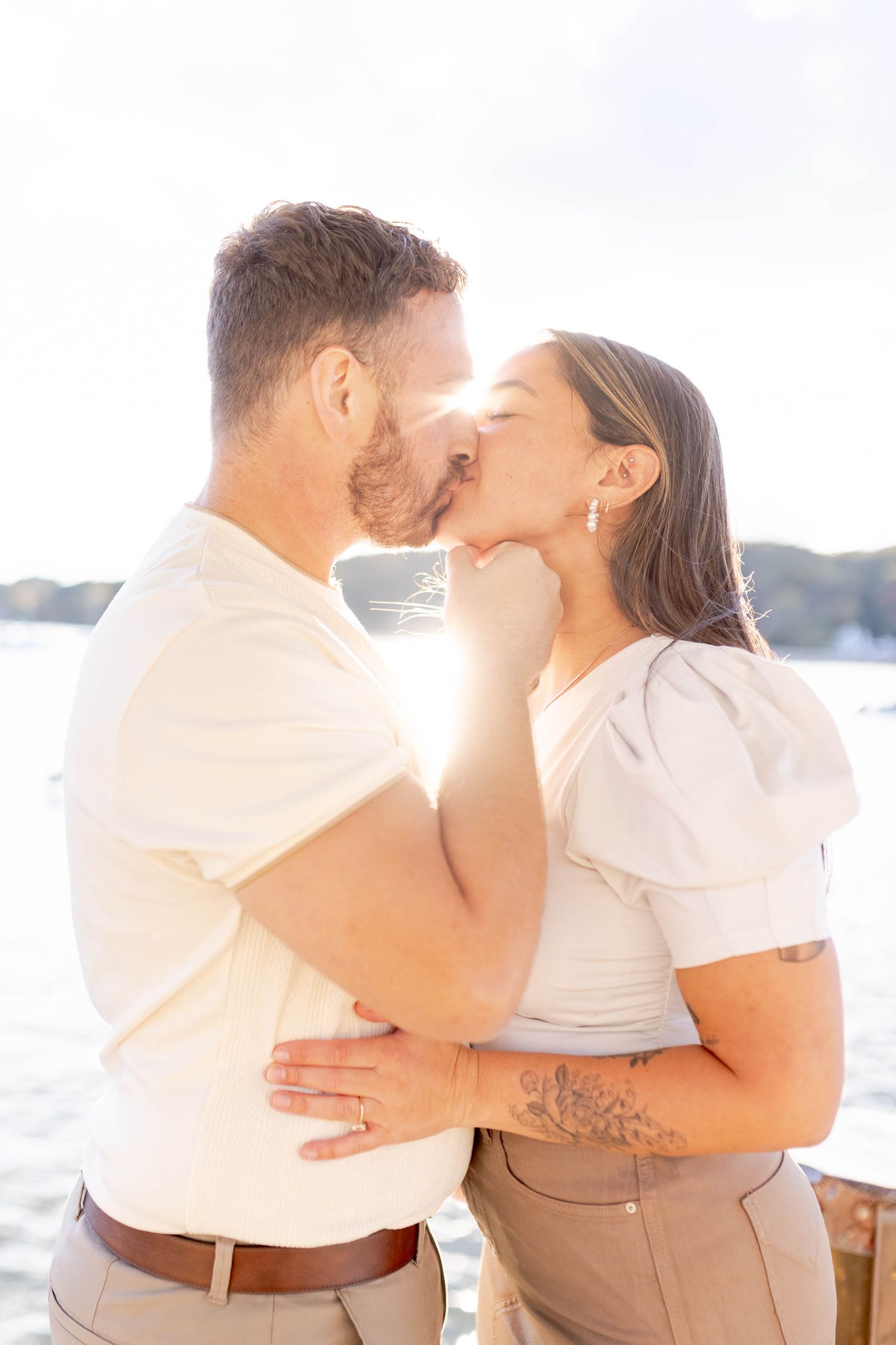 A romantic couple in white and beige outfits share intimate kisses by the water during sunset.