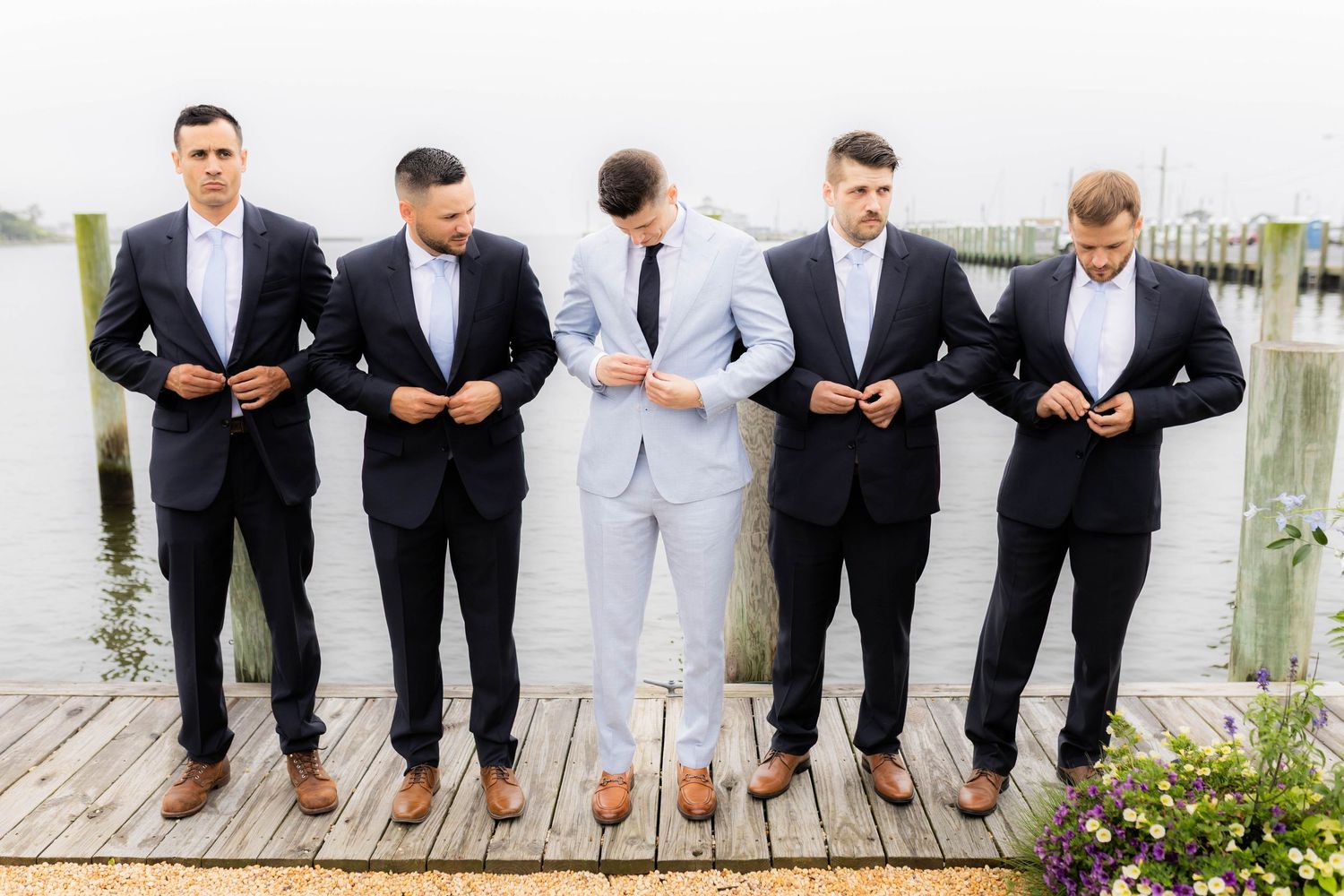 Groomsmen in formal suits showing off their colorful socks while standing on a dock by the water.