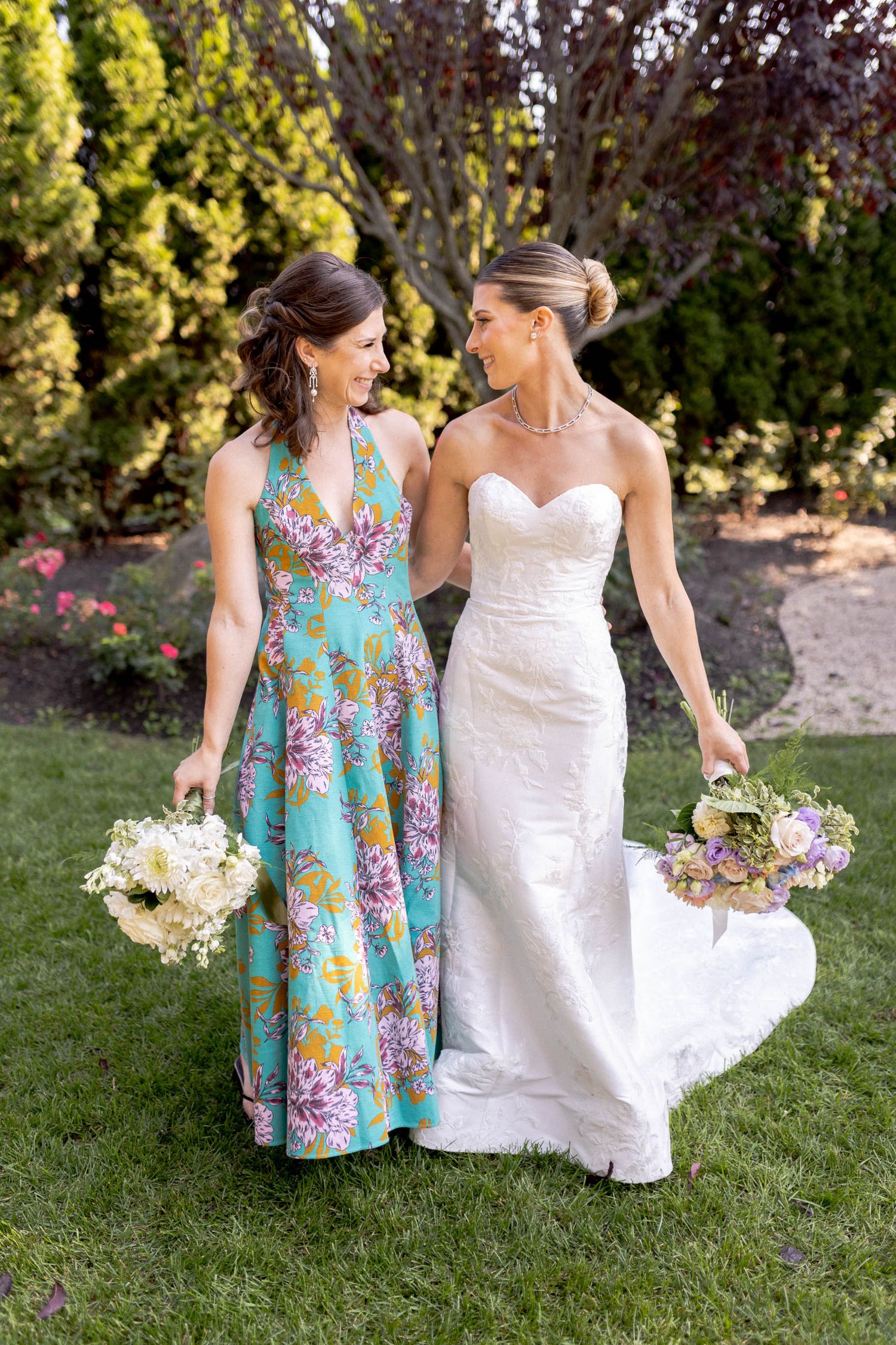 Two women in formal dresses walk together on grass, one wearing floral print and one in white, both holding flower bouquets.
