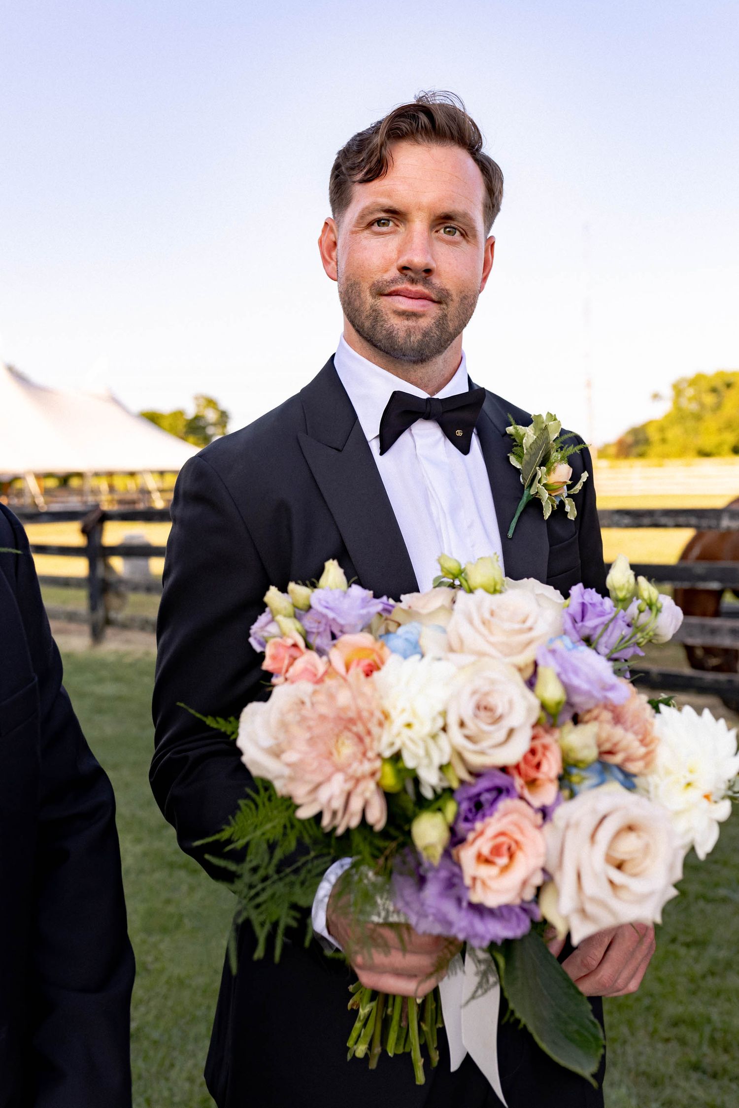 A person in a black tuxedo and bow tie holds a beautiful bouquet of pink, peach and purple flowers outdoors.