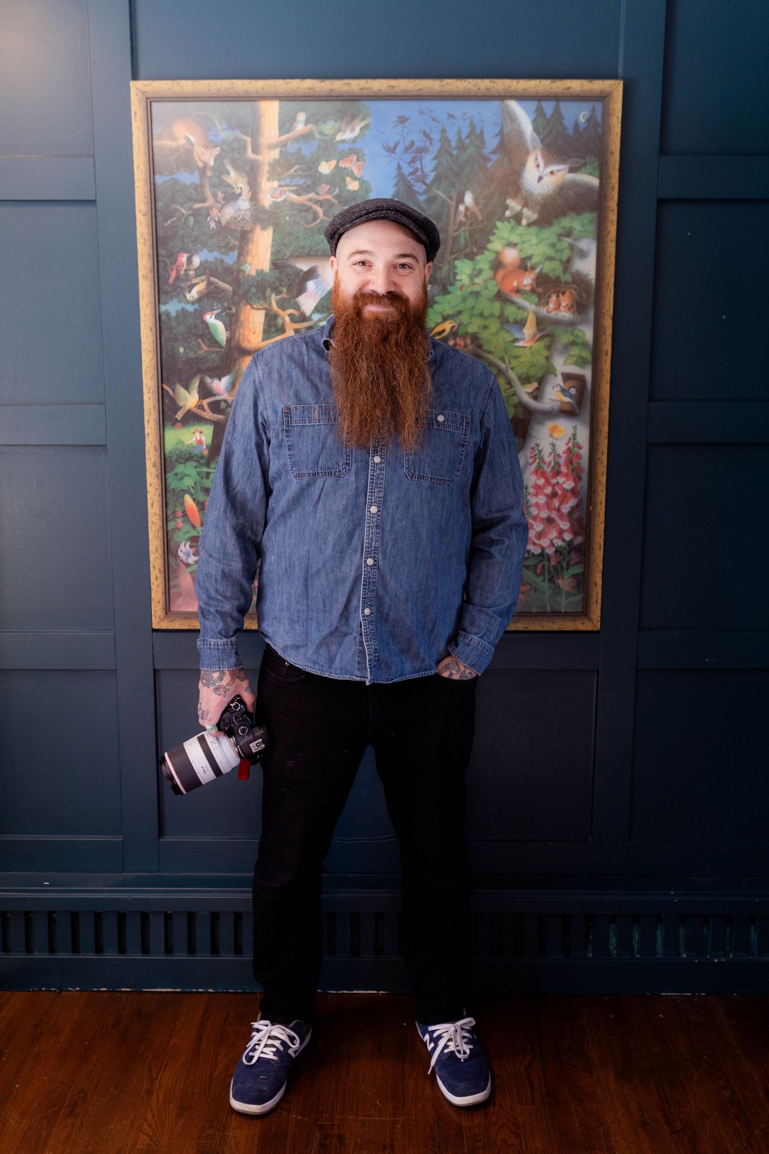 Bearded photographer in denim shirt poses with camera against dark wall with framed artwork. Steve Coiro. Fireside Sparrow Photography