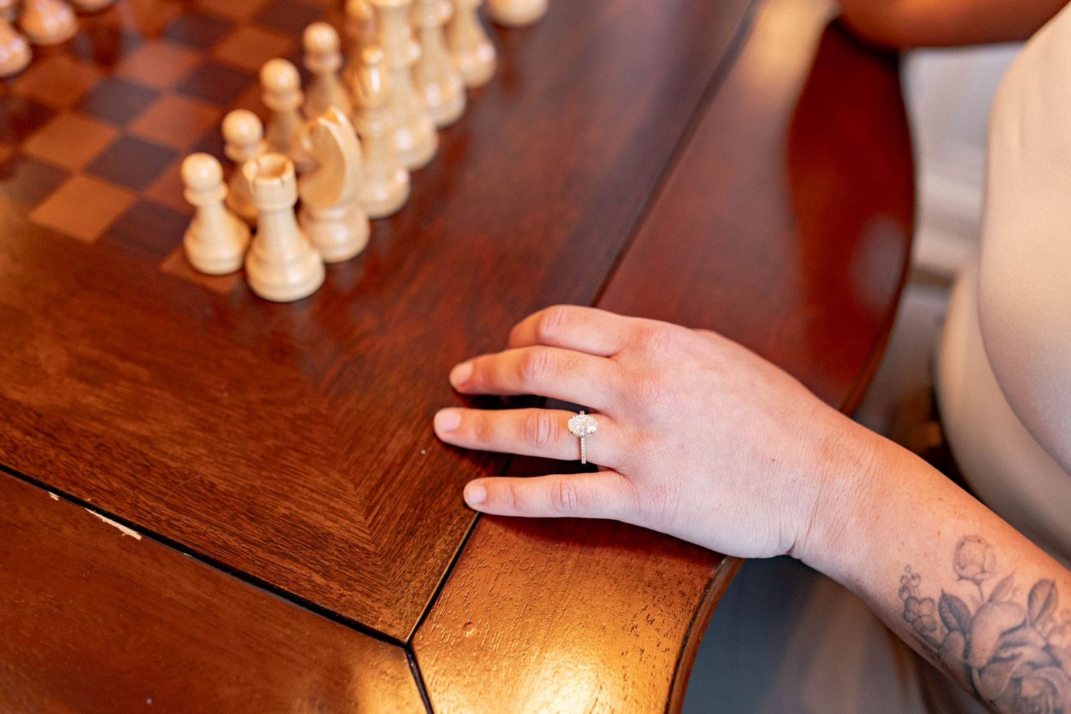 Hand with a ring and tattoo rests on a wooden chess board with white pieces set up for play.