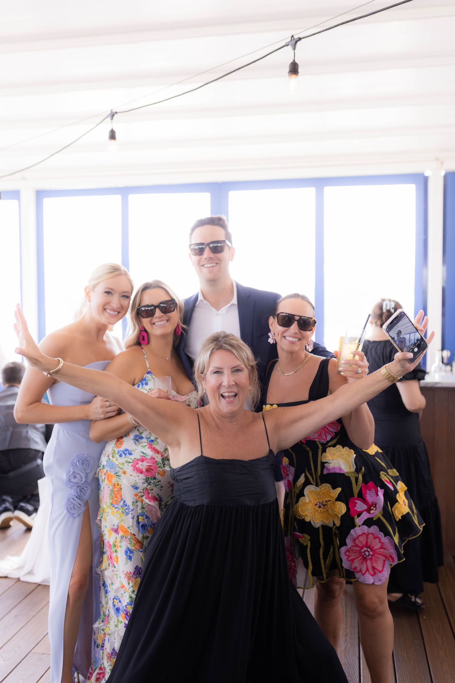 A group of wedding guests in summer attire pose together on a deck with their arms raised in celebration.