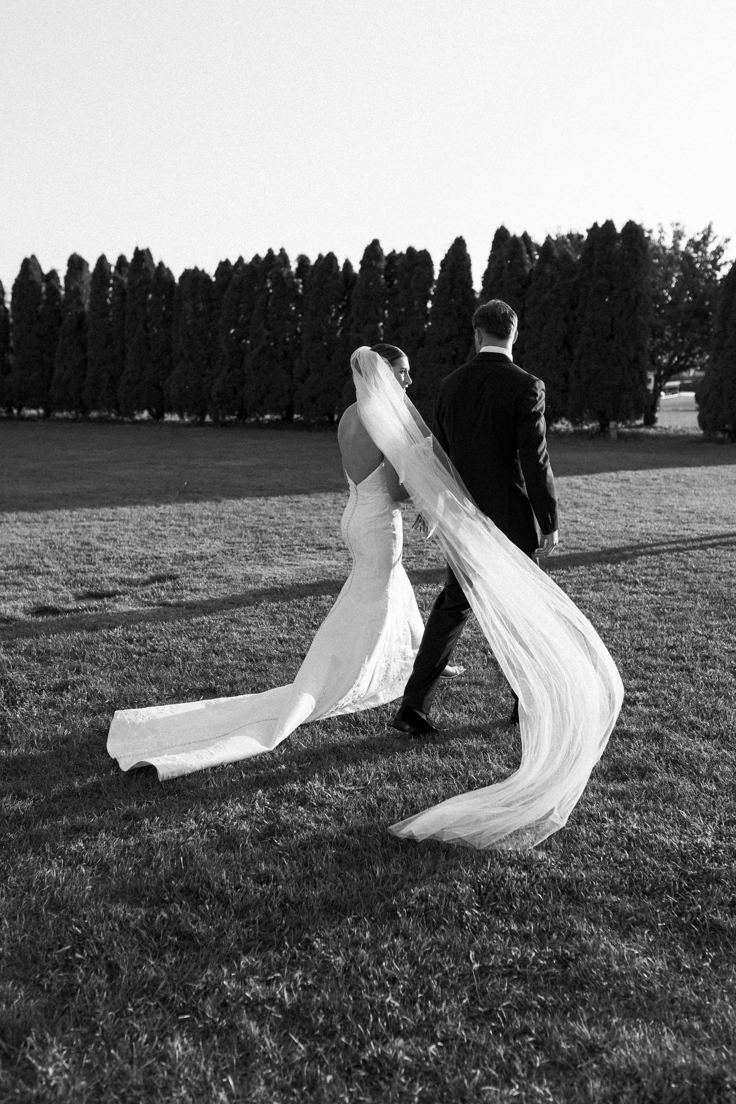 Black and white photo of a wedding couple with a flowing veil in a garden setting.