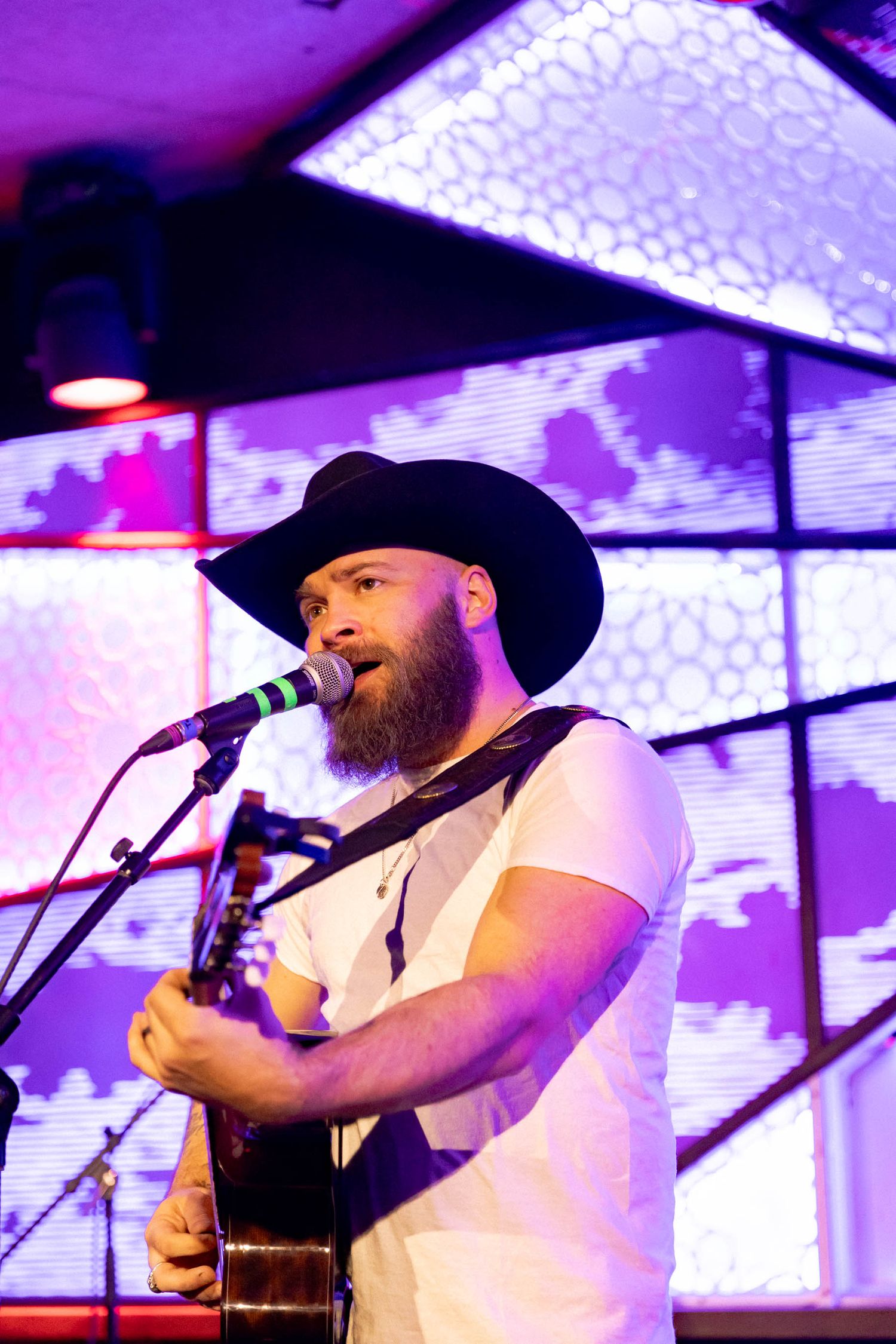 Musician in cowboy hat performs with acoustic guitar against geometric patterned backdrop.