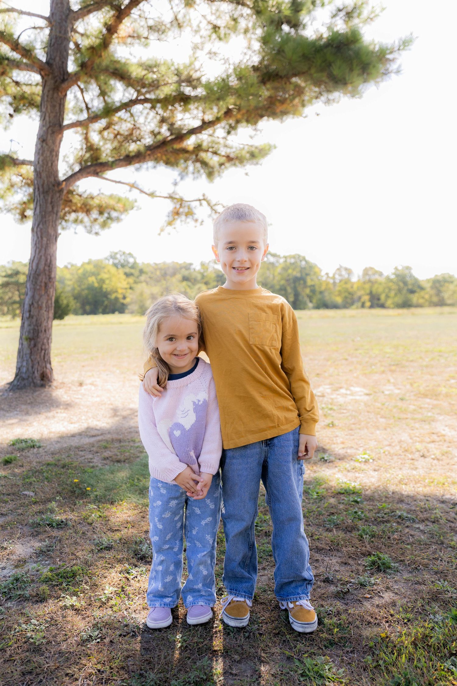 Two young children in casual clothes pose together under tall pine trees.