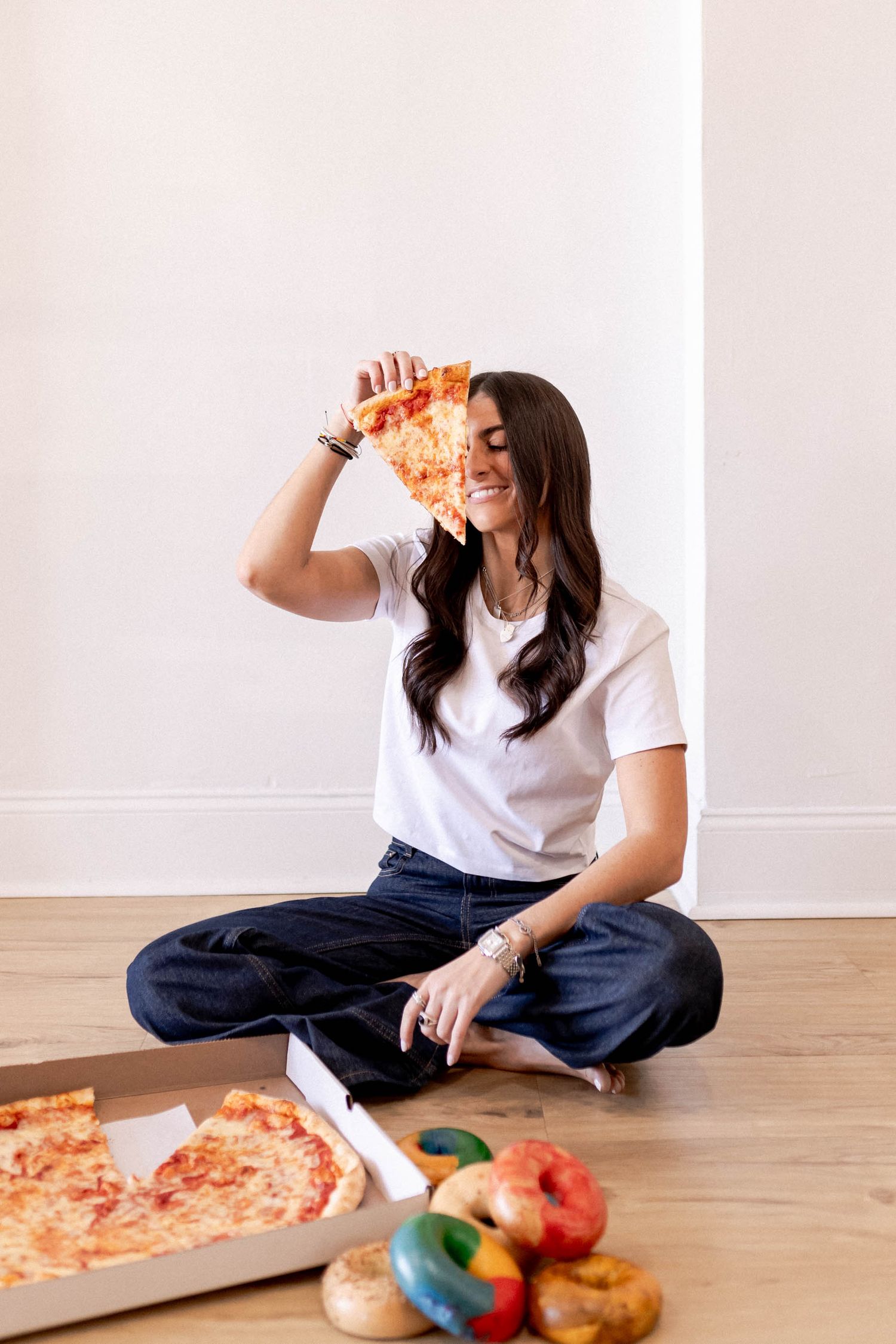 Person enjoying pizza while sitting cross-legged on wooden floor with colorful produce nearby.