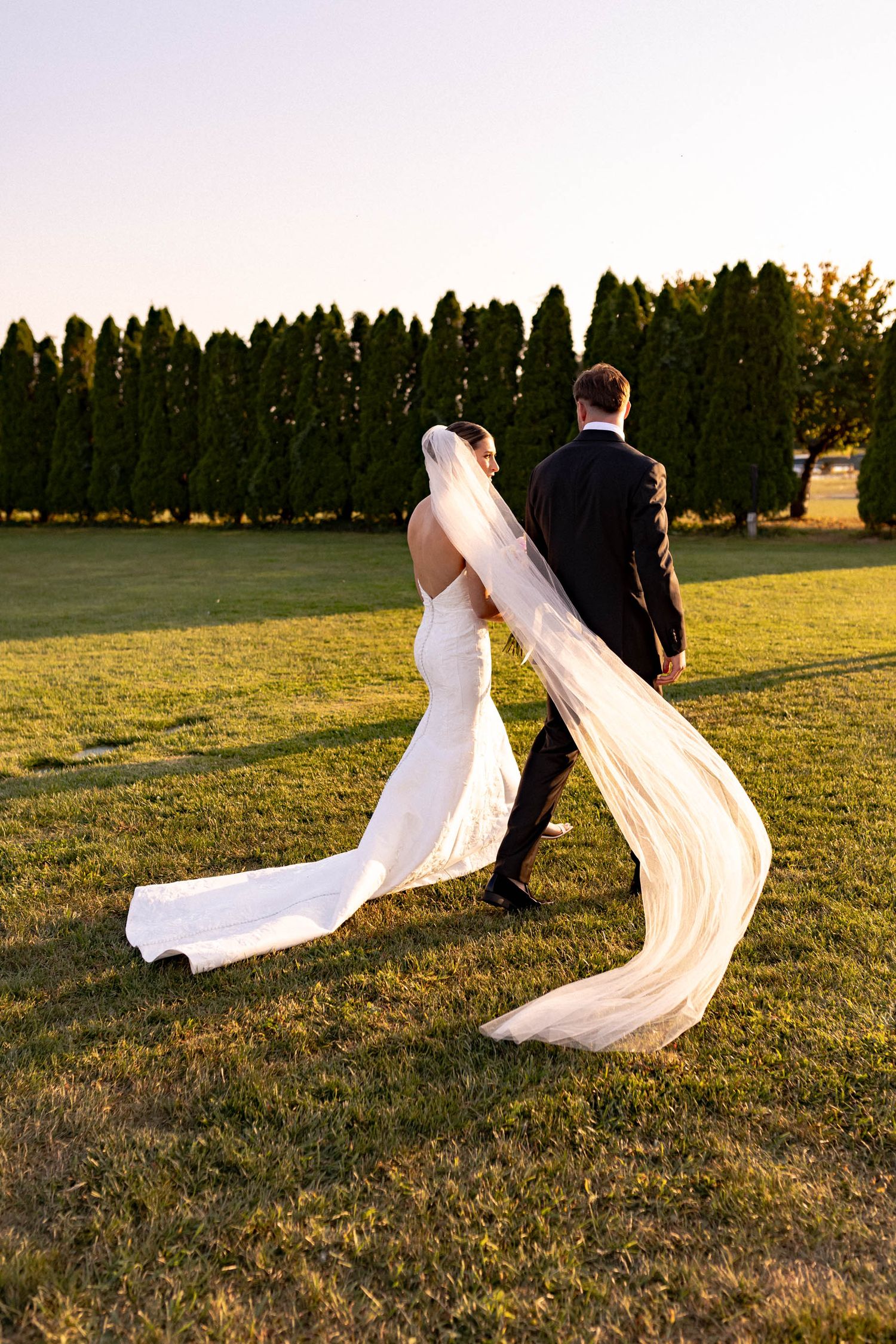Bride's flowing veil catches the golden sunlight as couple walks across field at sunset.