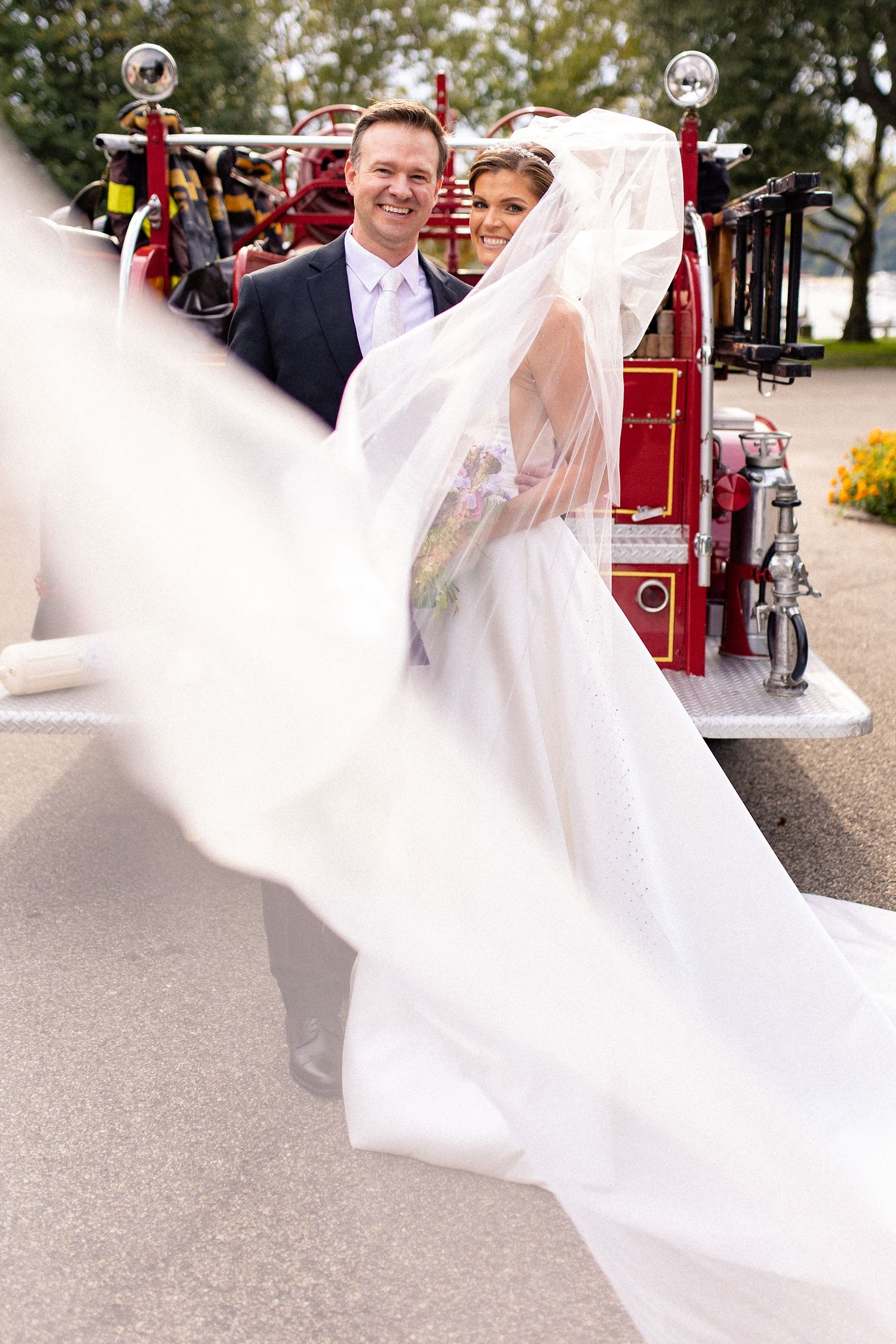 A wedding couple poses in front of a vintage red fire truck with a flowing white veil caught in the breeze.