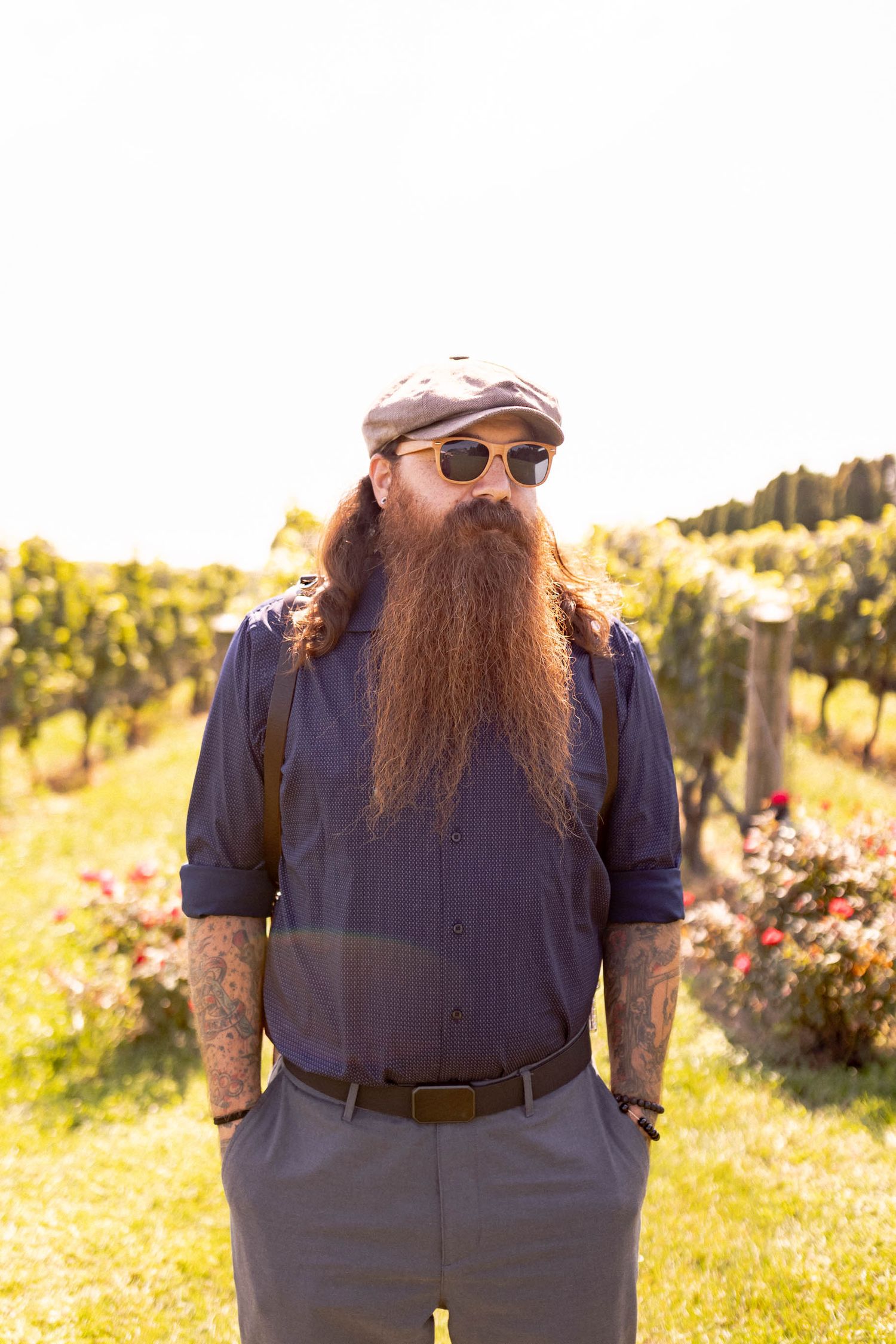 A bearded person wearing suspenders and sunglasses stands in a sunny field with wildflowers and trees in the background.