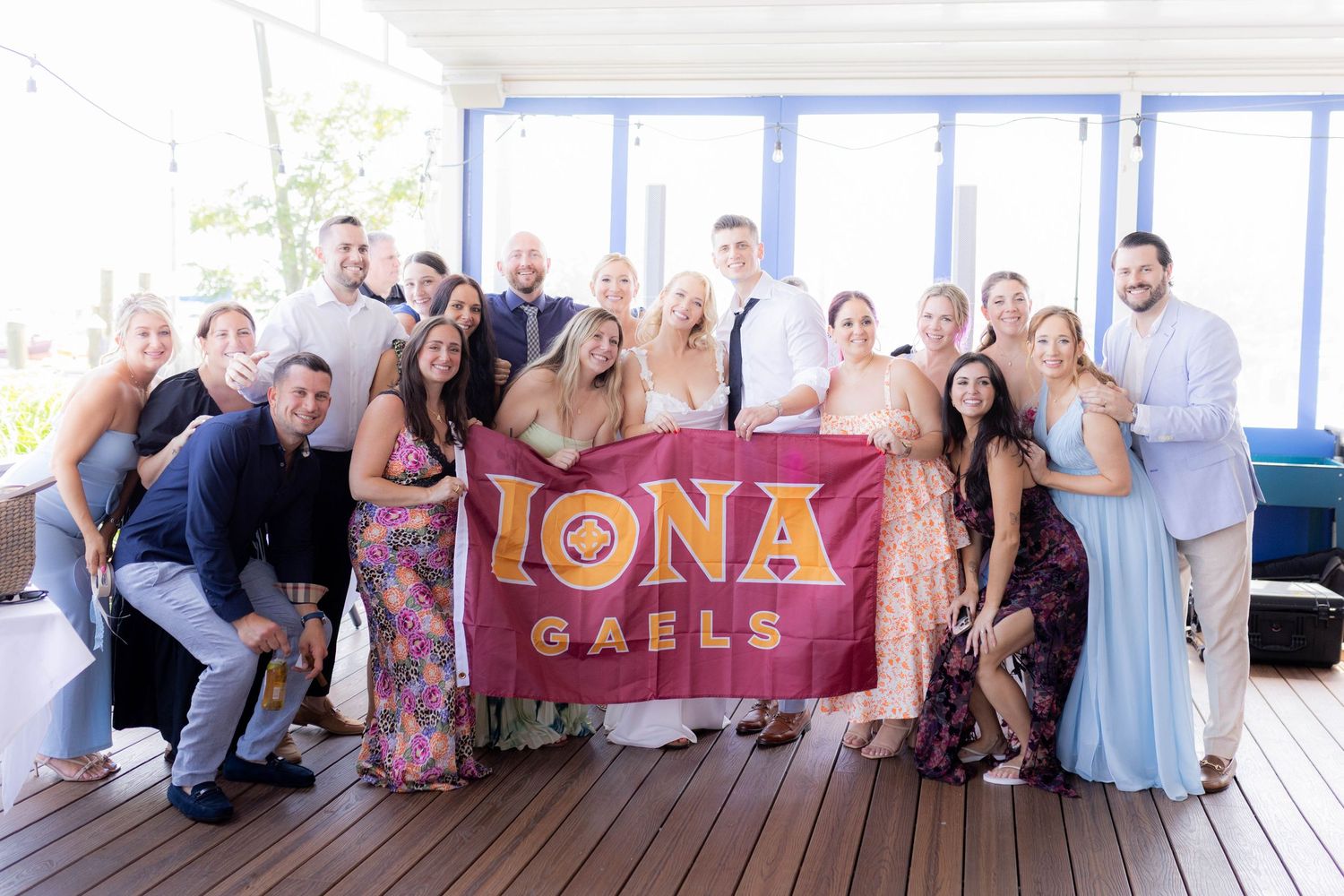 Wedding guests gather for a panoramic group photo on a deck with an Iona banner visible in the background.