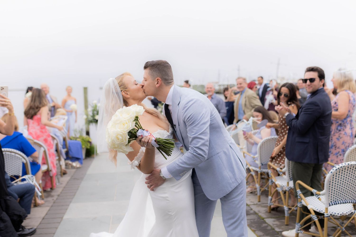 A couple shares a romantic kiss at their outdoor beach wedding ceremony as guests look on and celebrate.