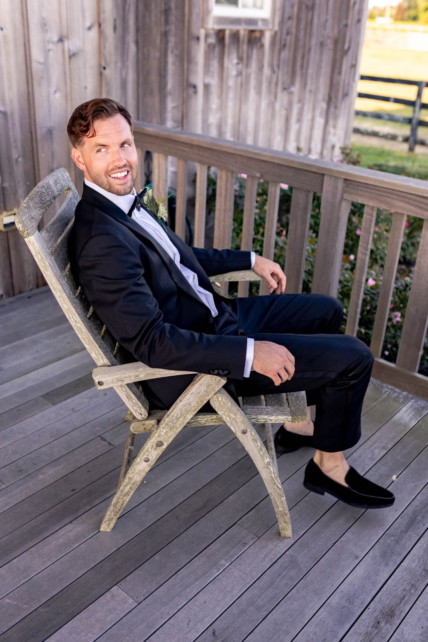 A person in a dark suit and tie relaxes in a wooden deck chair while smiling at the camera.