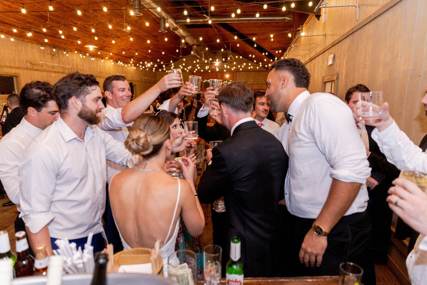 Wedding reception party guests dancing and celebrating under warm string lights in a rustic venue.