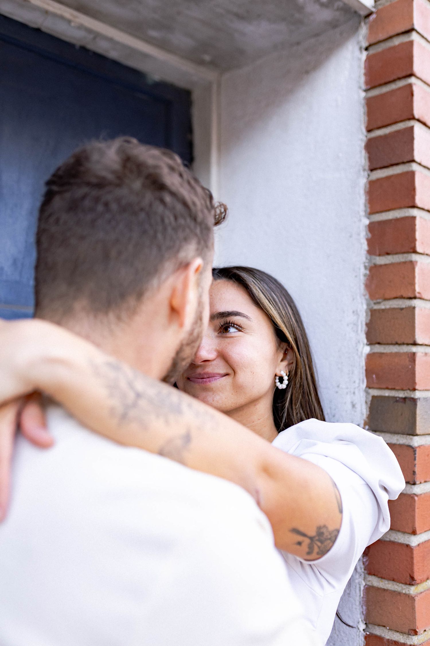 A romantic couple shares an intimate moment against a white wall with brick columns.