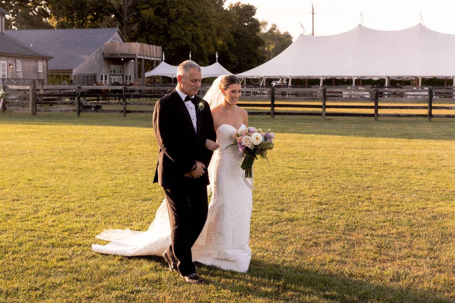 A couple walks together on a grassy field at sunset with white tents and fencing in the background.