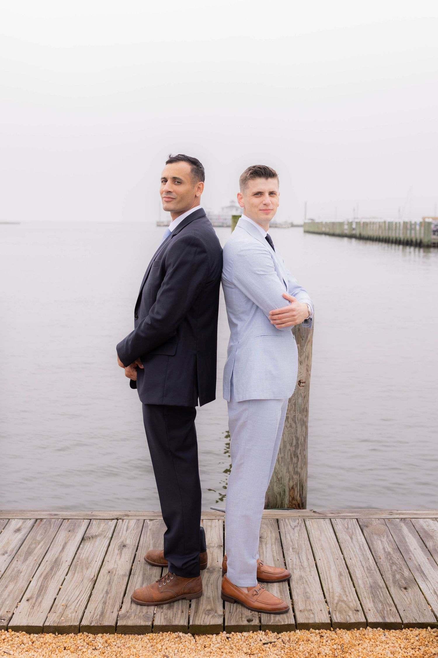 Two people in contrasting suits stand back-to-back on a wooden pier overlooking a misty waterfront.