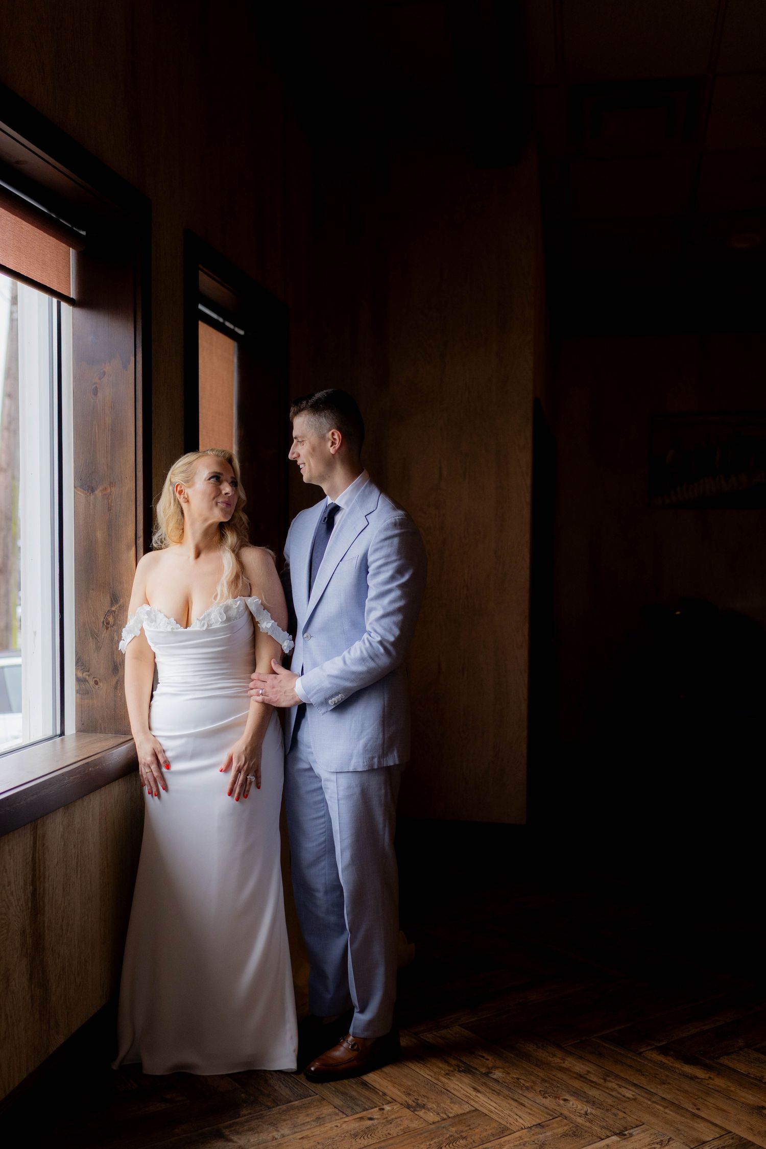 Couple in formal attire standing by window in dark rustic room.