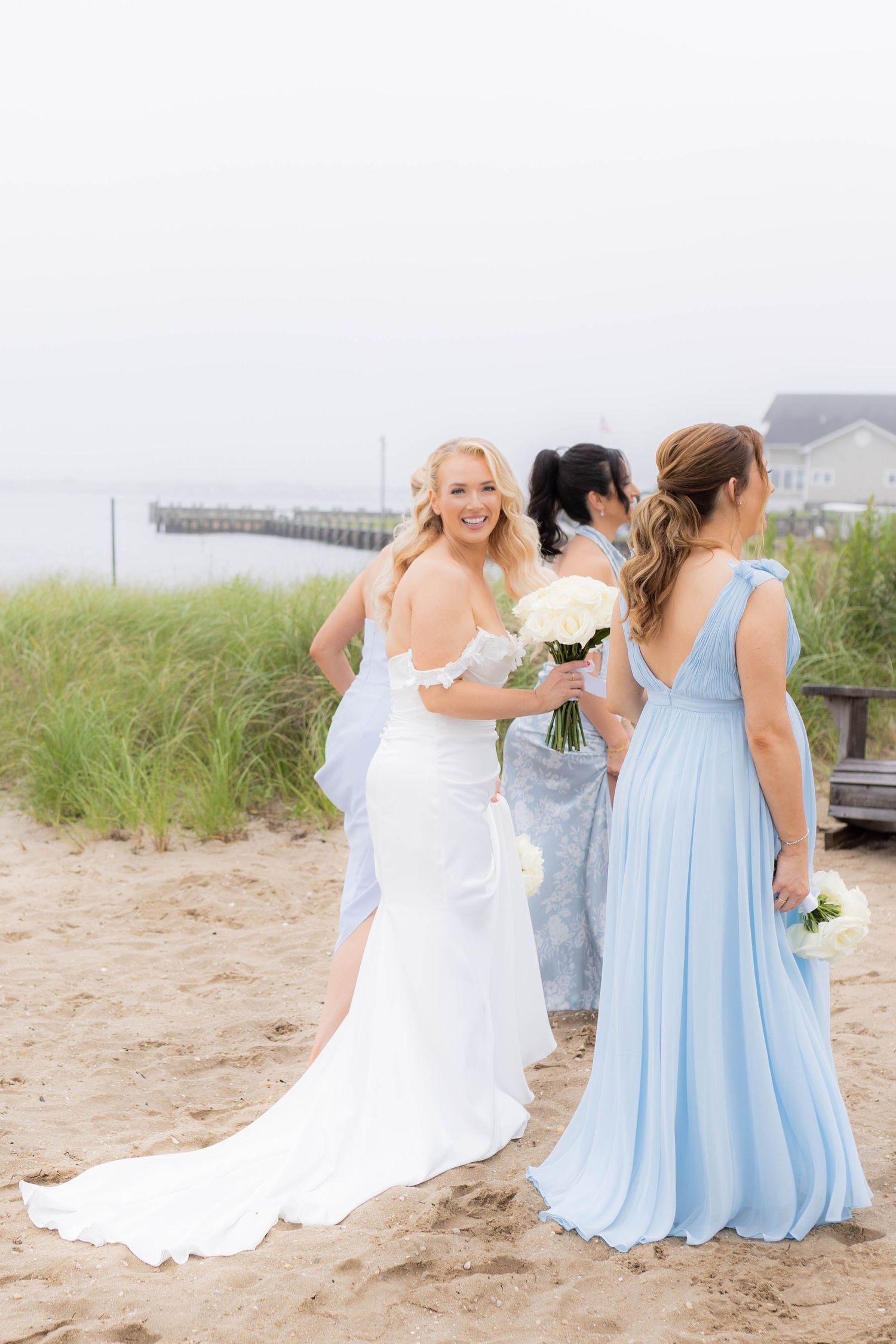 A wedding party in white and light blue dresses poses on a sandy beach with dunes and a pier in the background.