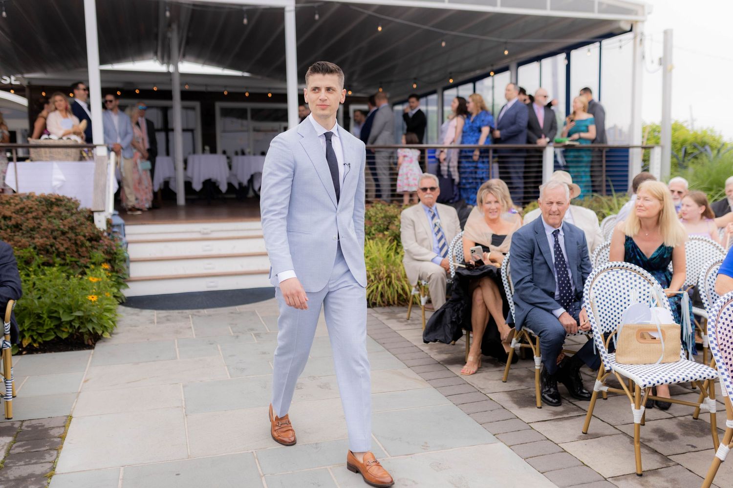 Wedding guests gather outside on a sunny day at a venue with a covered walkway and seating area.