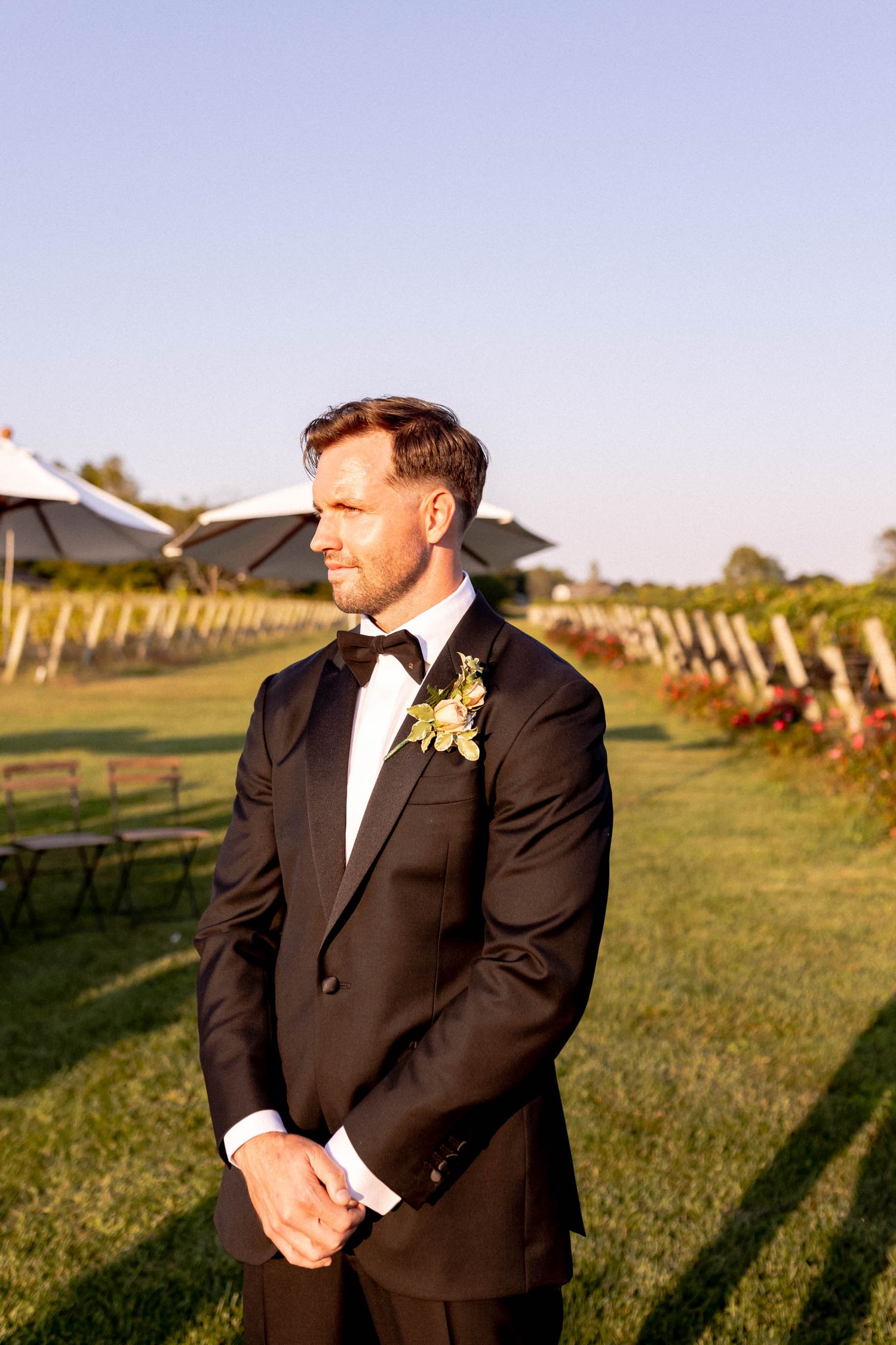 A groom in a classic black tuxedo and bowtie stands in front of a vineyard at sunset.