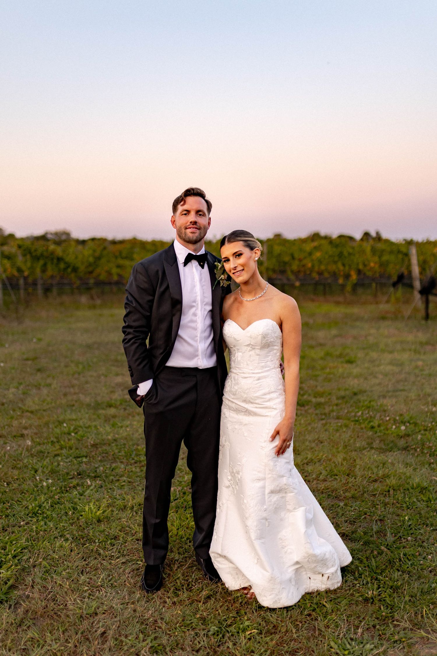 A couple poses together in formal wedding attire at sunset in a vineyard.
