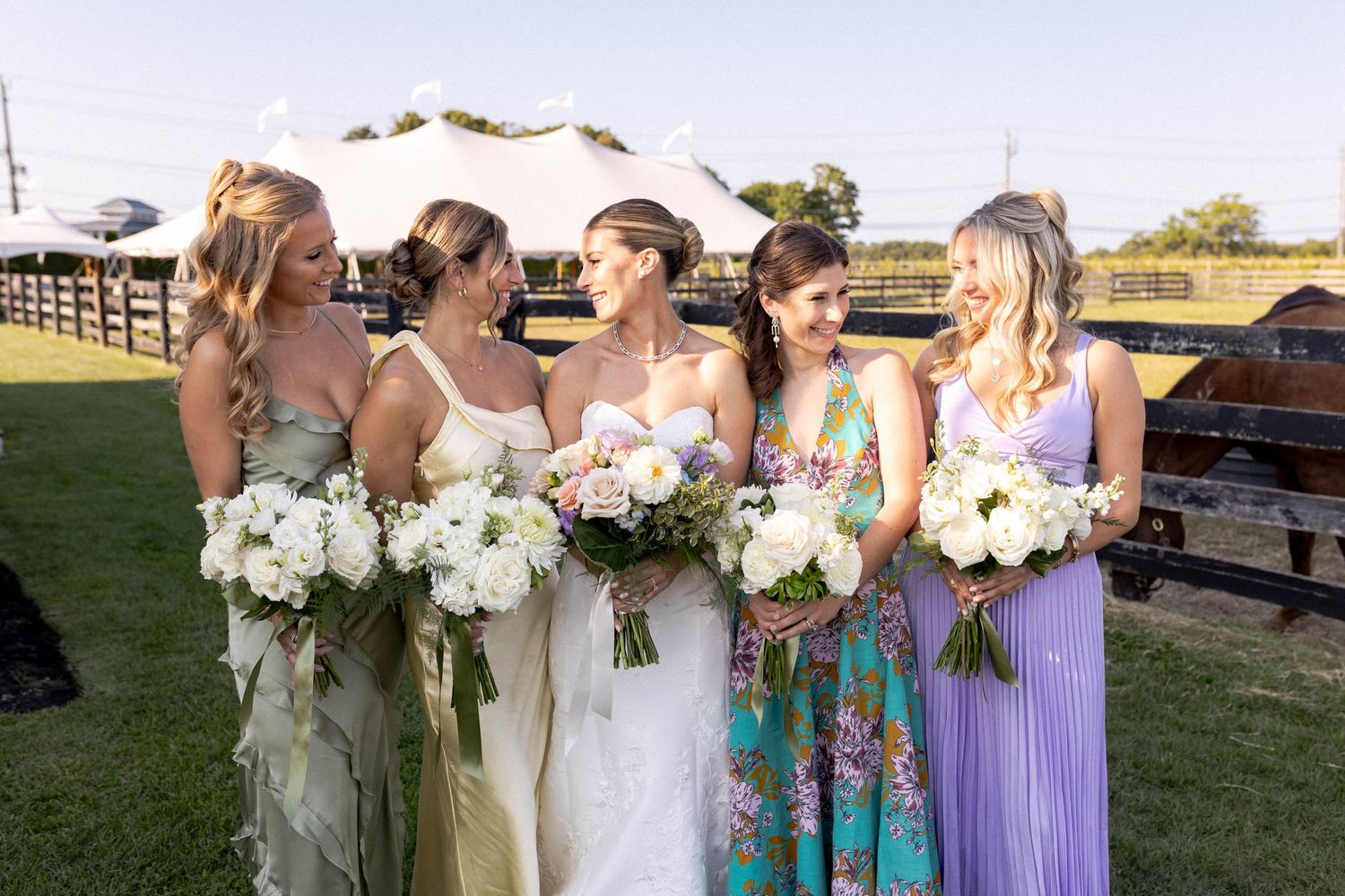 Wedding party members hold white floral bouquets while standing in a row on a grassy lawn at sunset.