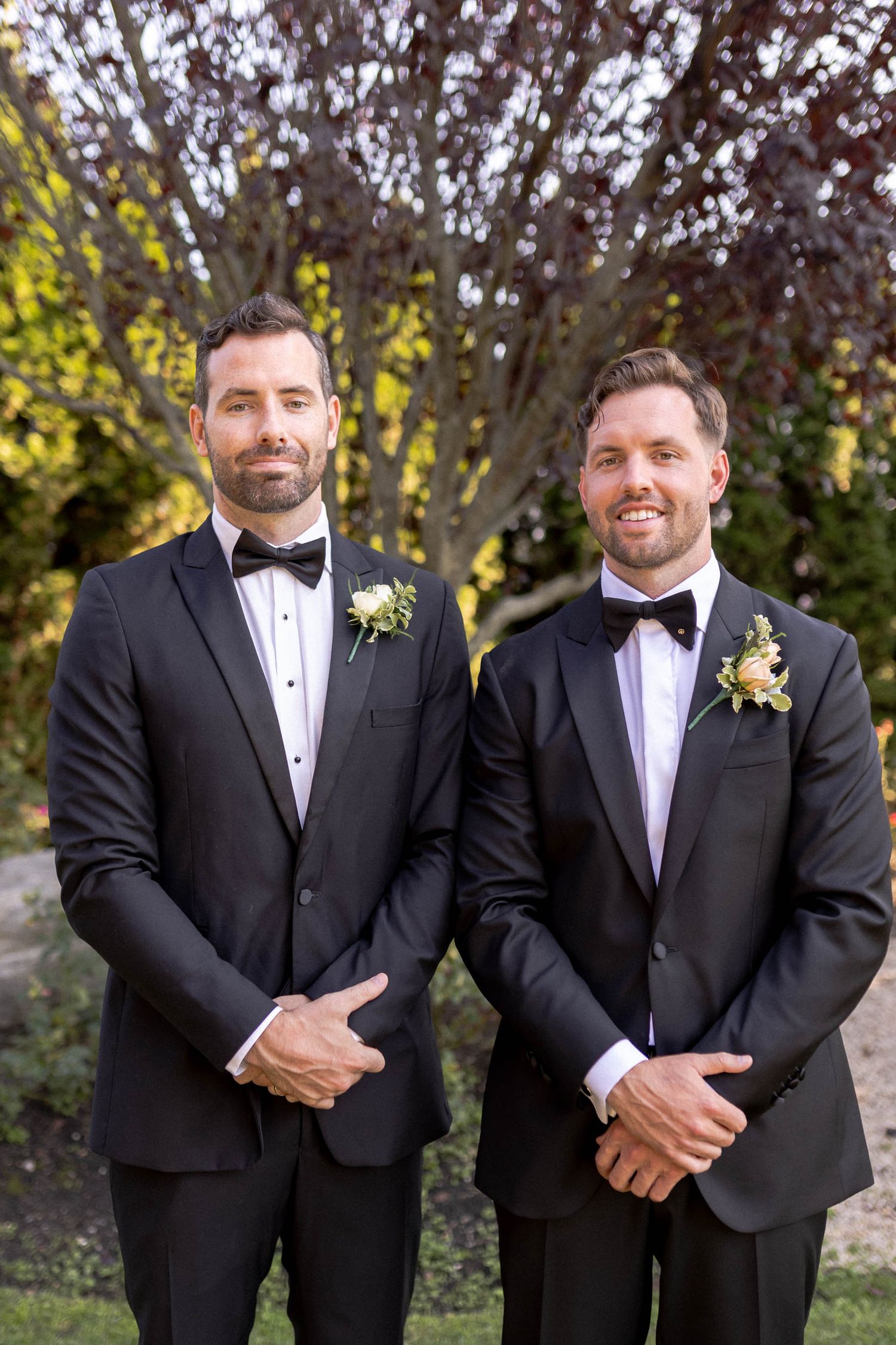 Two people in black tuxedos and bow ties stand together outdoors with boutonnières on their lapels.