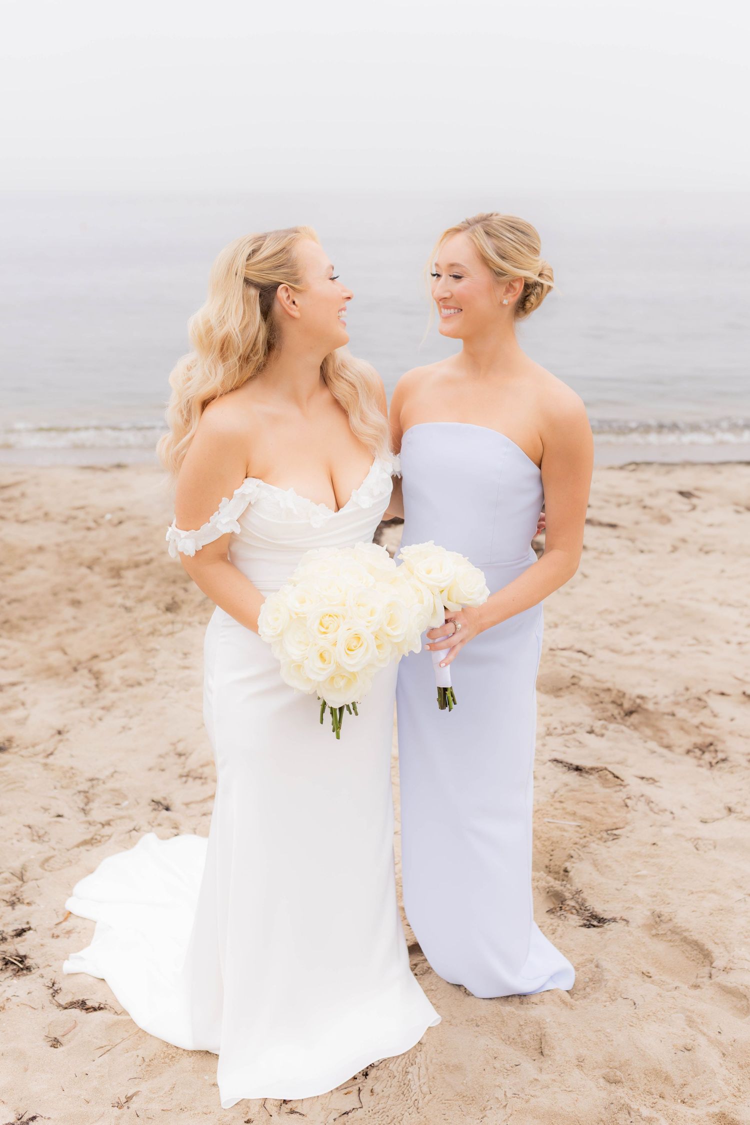 Two people in white dresses share a joyful moment together on a sandy beach with ocean waves in the background.