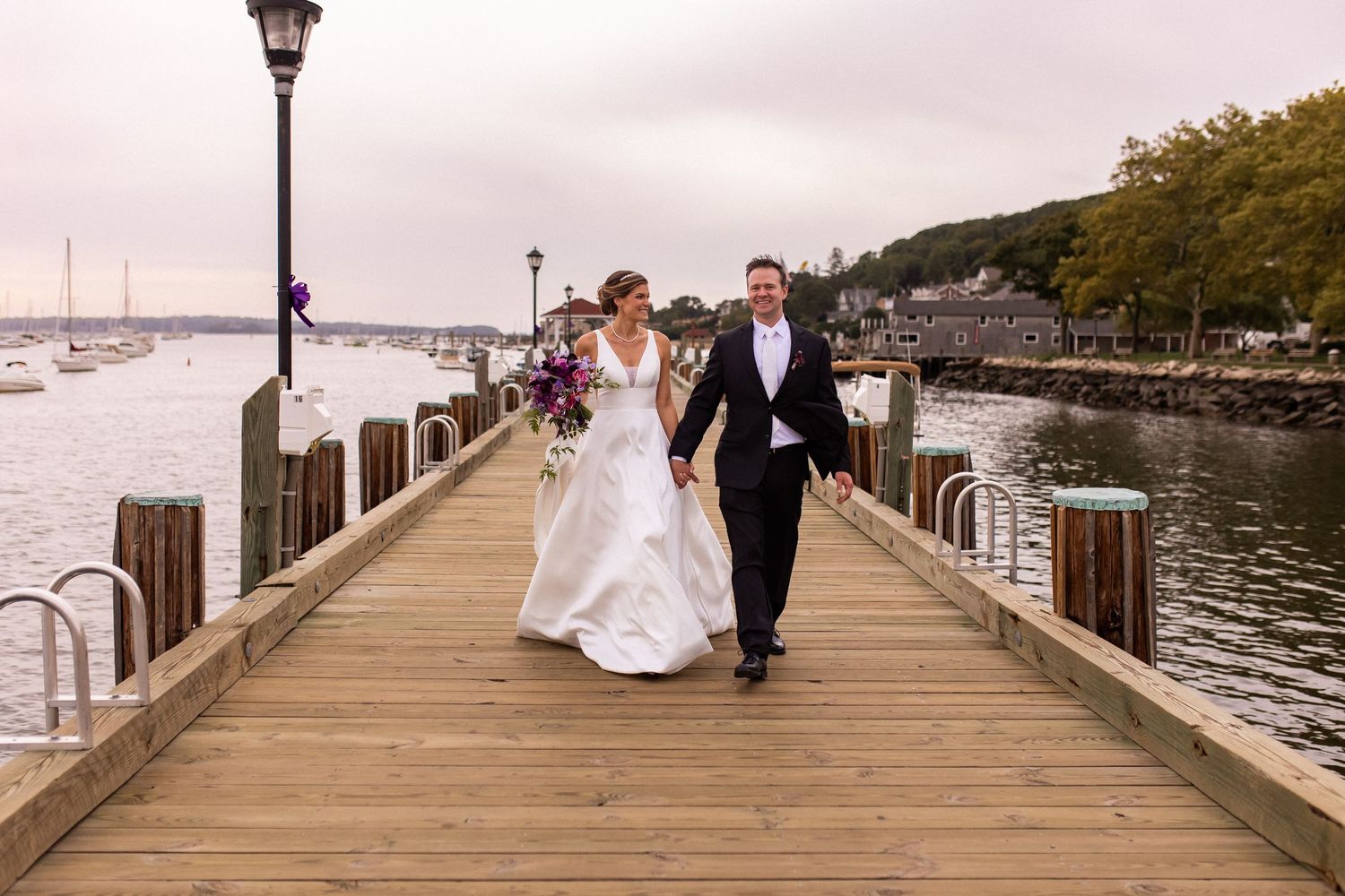 Wedding couple walks along a scenic marina boardwalk with boats and water in the background.