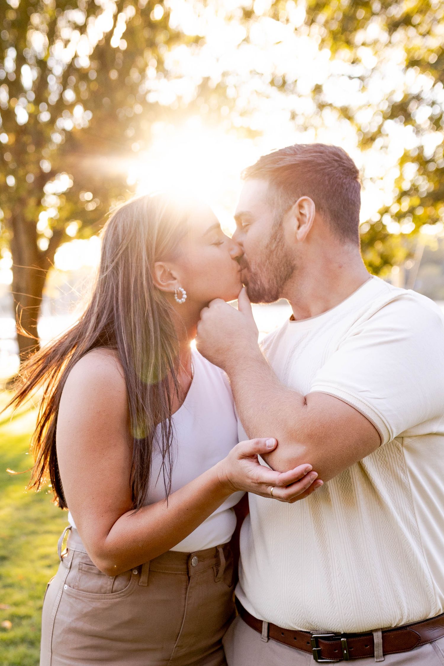 A romantic couple shares a tender kiss in a sun-drenched outdoor setting with golden light filtering through trees.